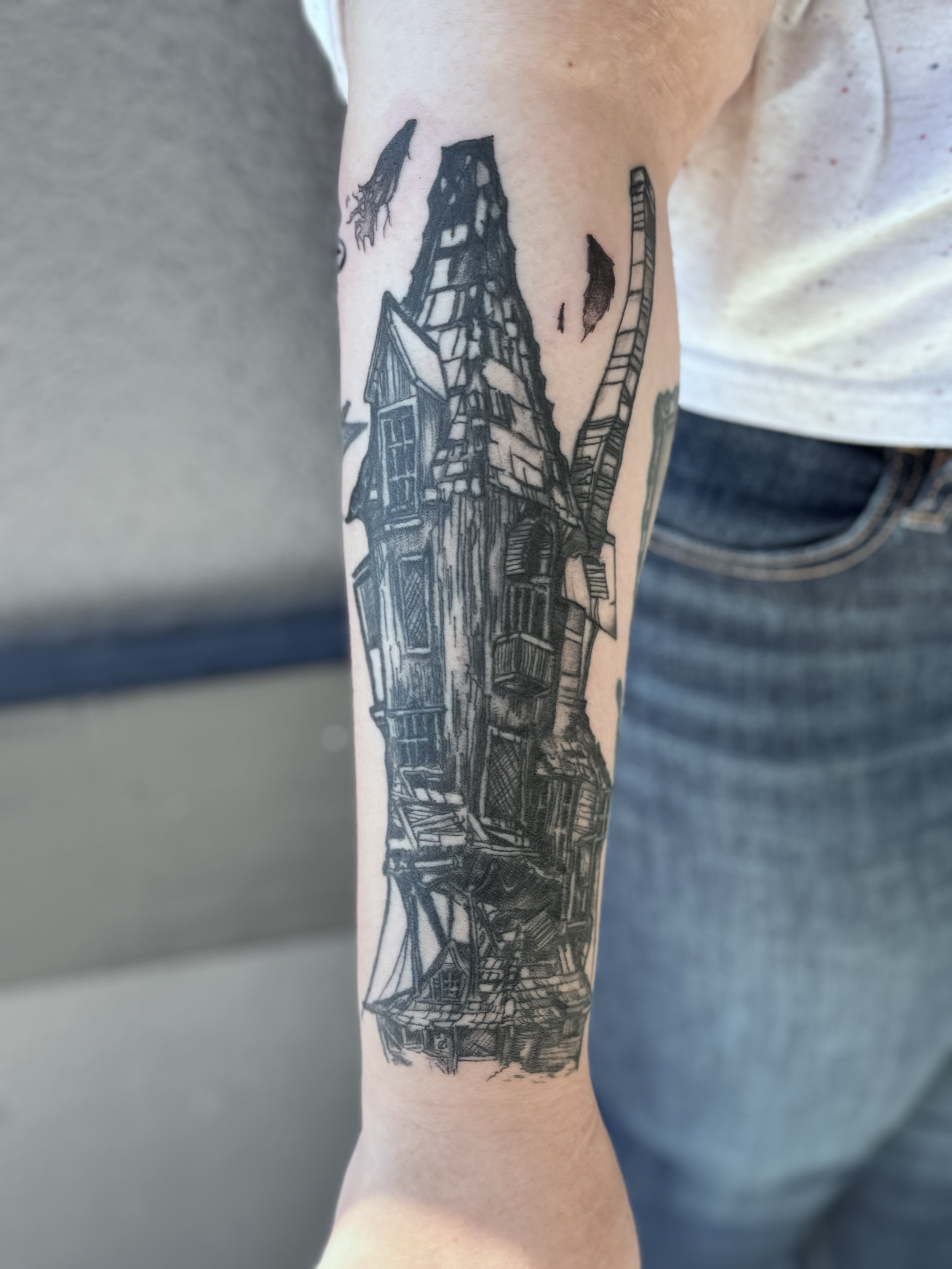 Black and gray tattoo of a whimsical, crooked house with multiple levels, turrets, and a leaning tower on a person's forearm, with a partly cloudy sky above.
