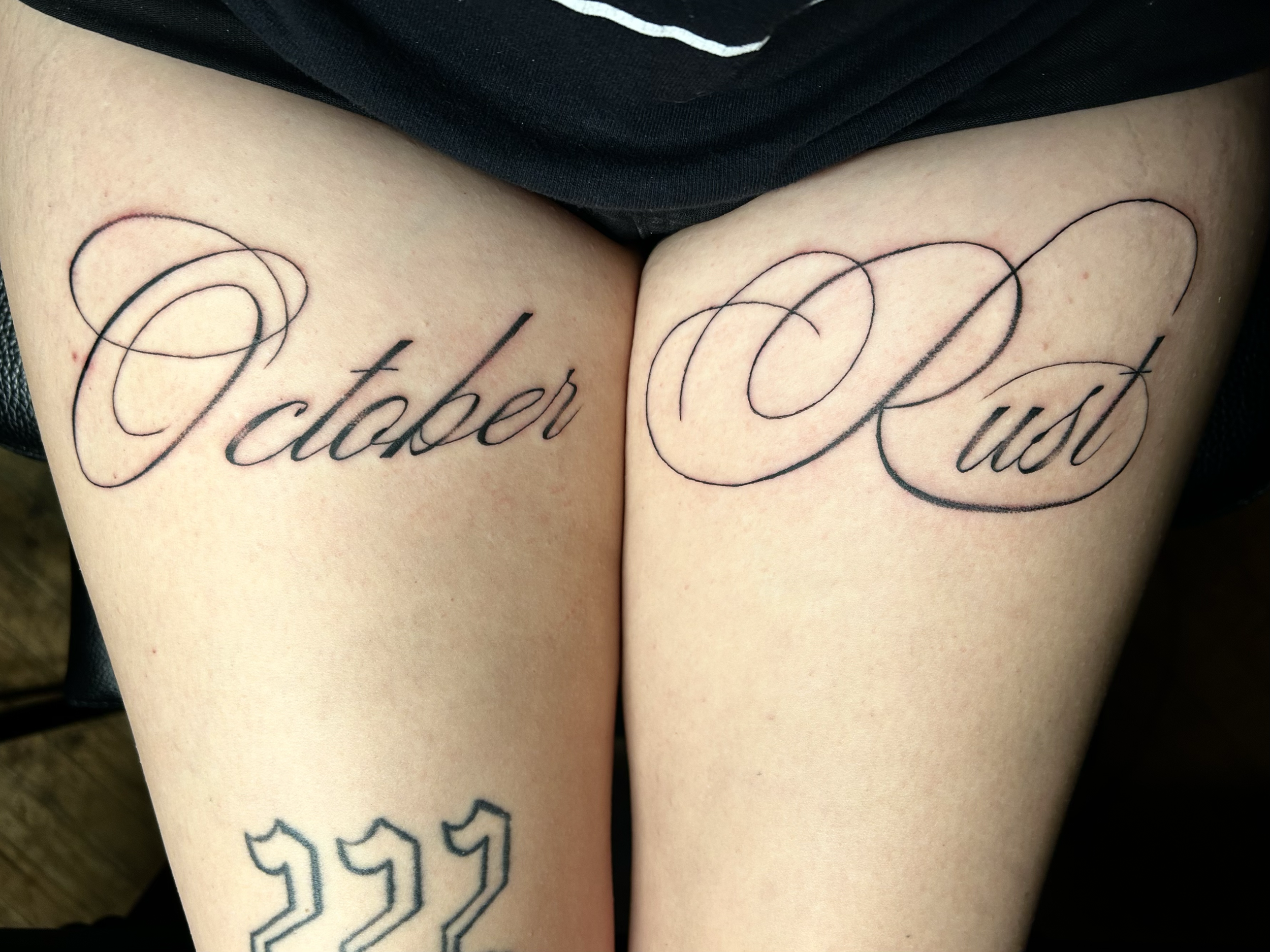Tattoo on thighs reading 'October' on the left thigh and 'Past' on the right thigh in elegant cursive script.
