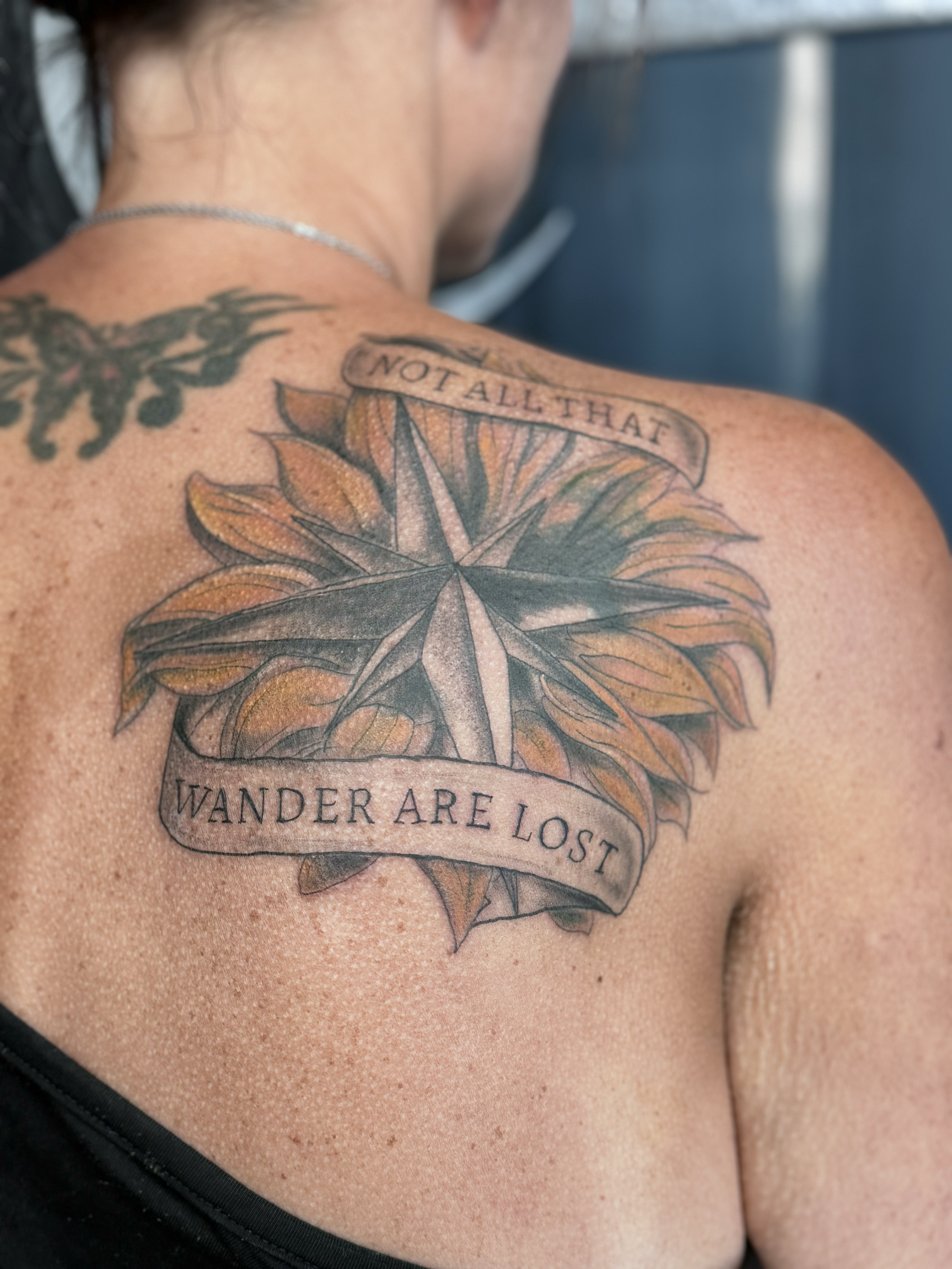 Tattoo on a person's shoulder featuring a large leaf with a banner that reads 'NOT ALL THAT WANDER ARE LOST.'