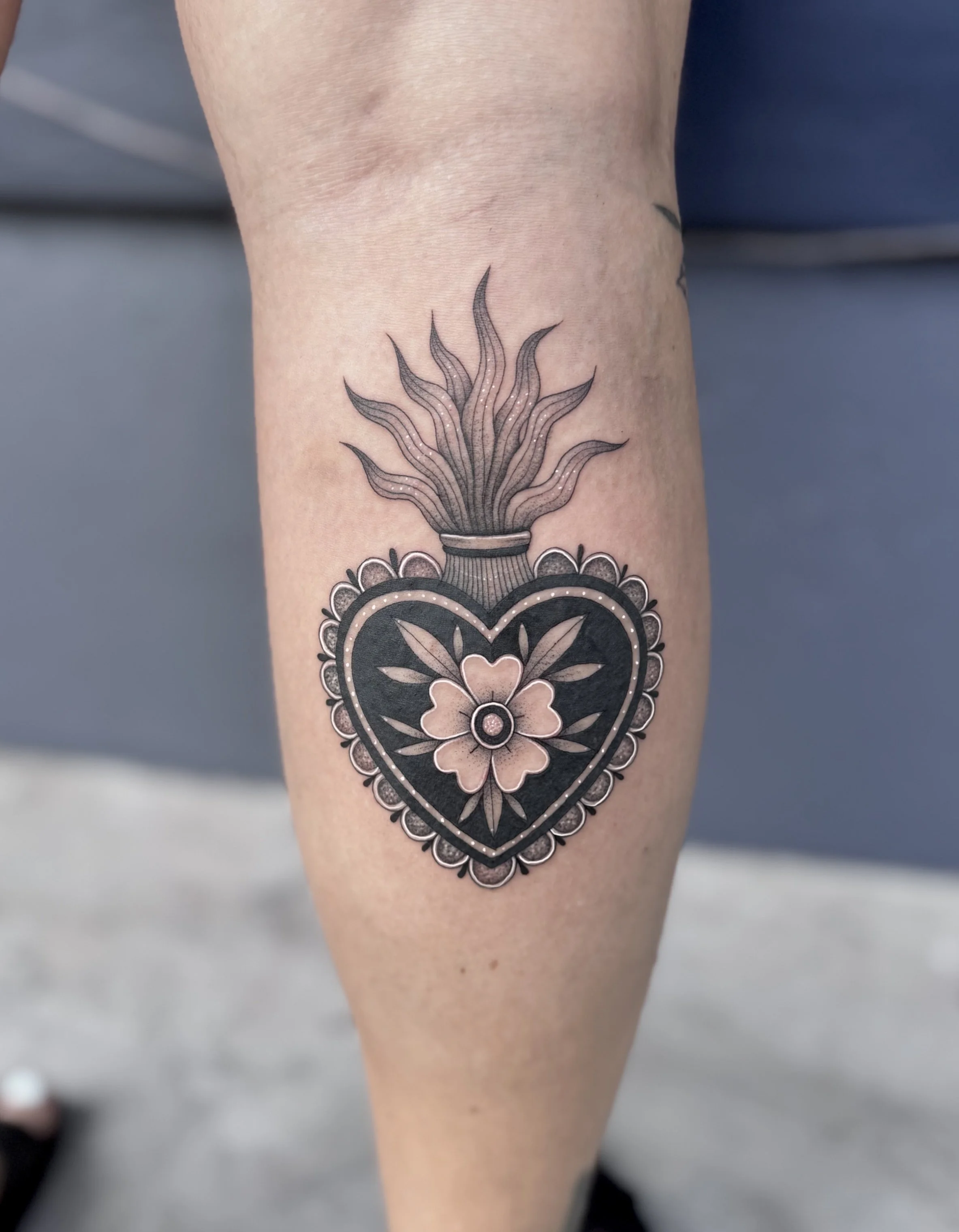 Tattoo of a heart-shaped floral design with a flower at the center, surrounded by a decorative border, and a flame-like element above.