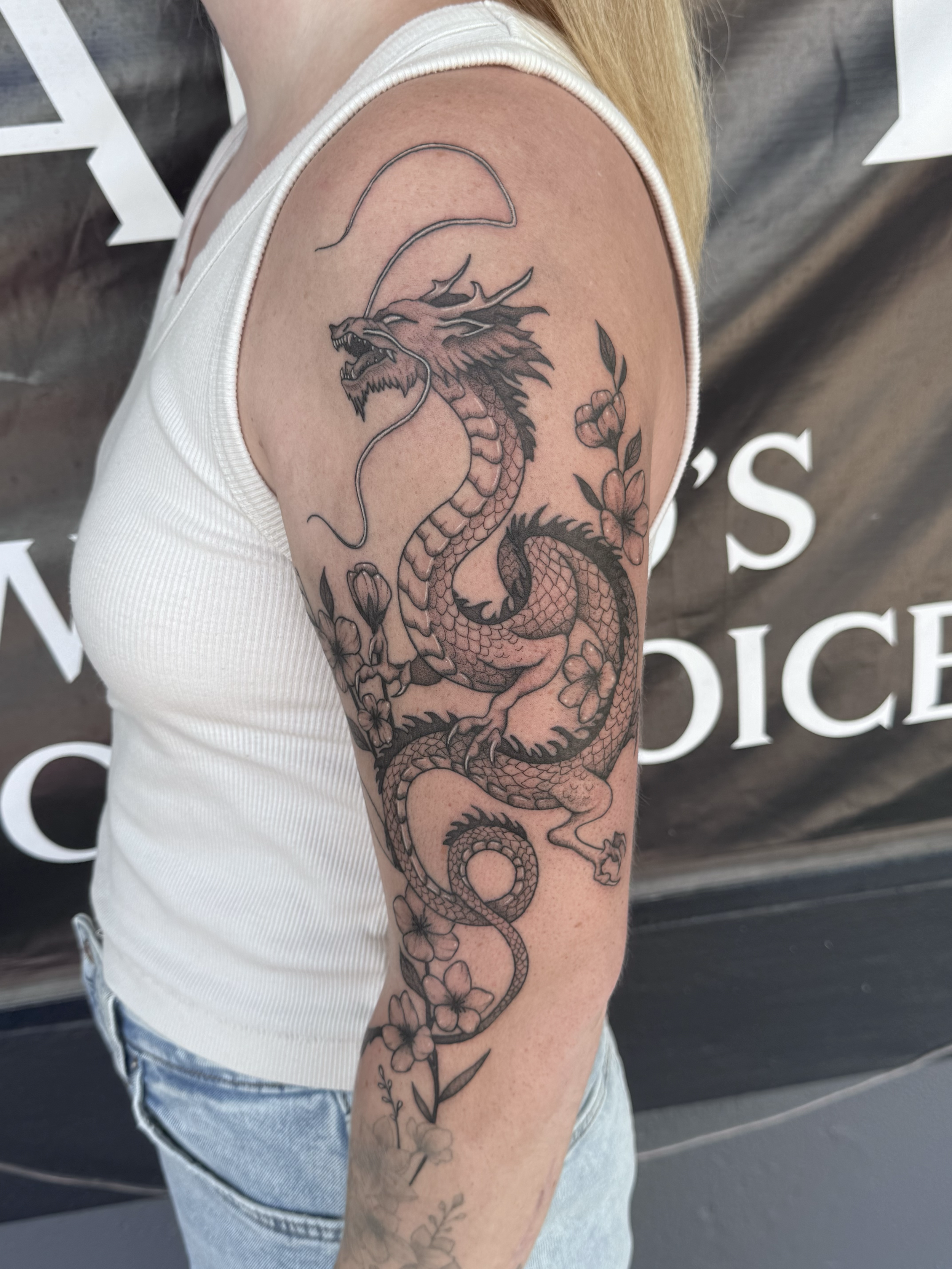 Black ink tattoo of a dragon surrounded by flowers on a woman's upper arm.