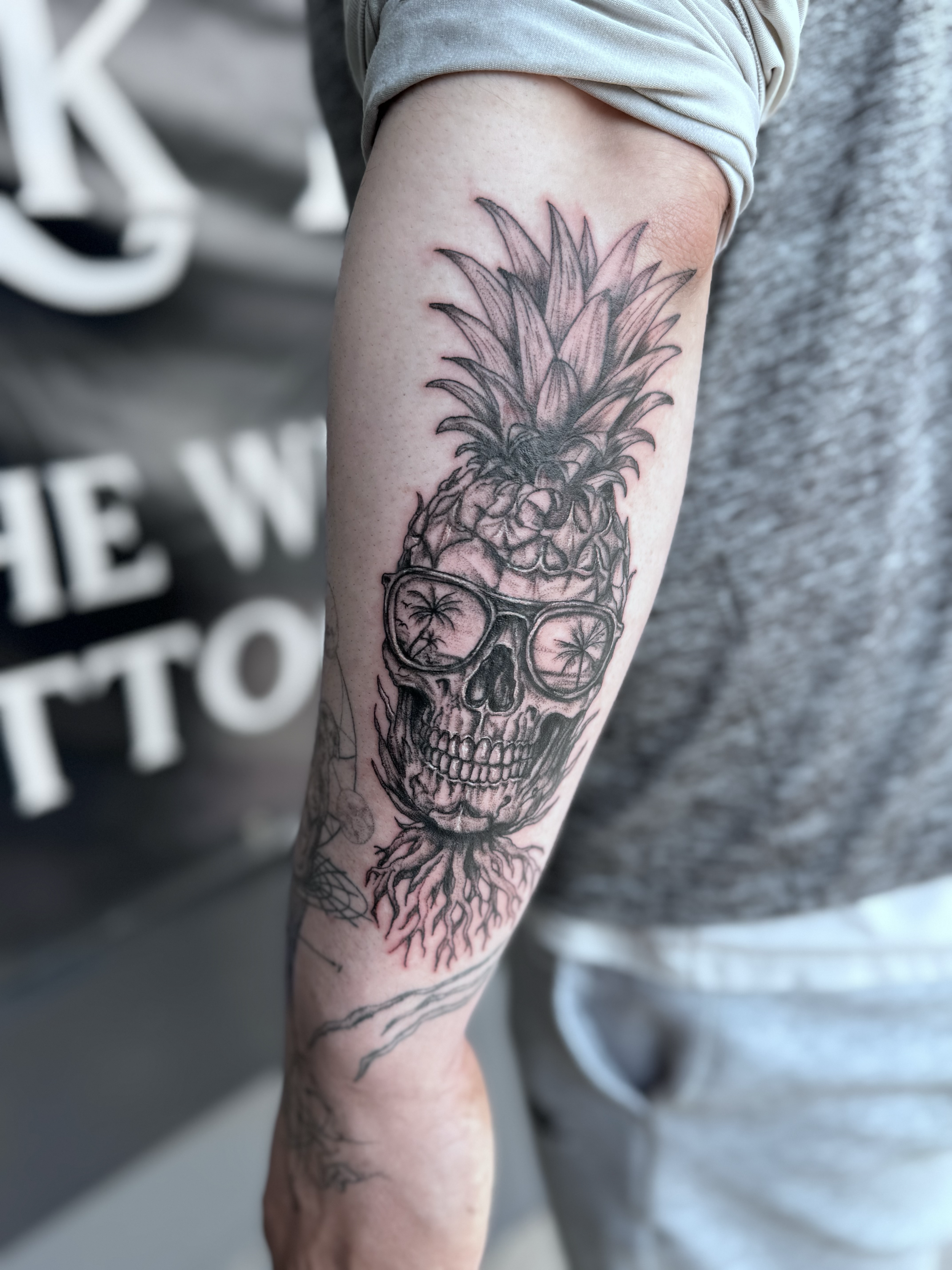 Tattoo of a skull wearing sunglasses, with a pineapple on top and roots at the bottom, on a person's arm.