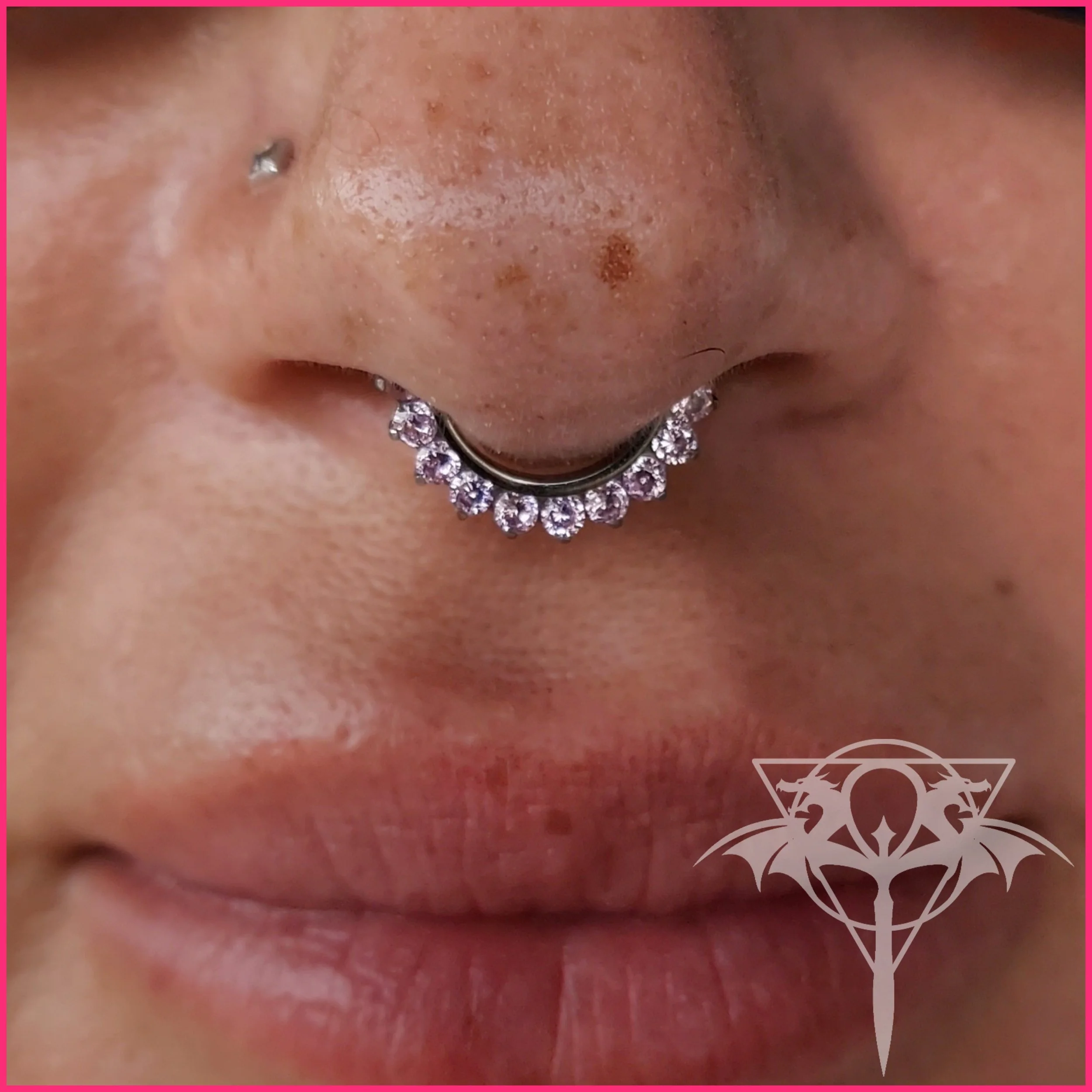 Close-up of a person's nose with a septum piercing decorated with small, clear rhinestones, and a lip piercing. The person's skin has freckles and a small mole. There is a pink border around the image and a stylized dragon logo in the bottom right co