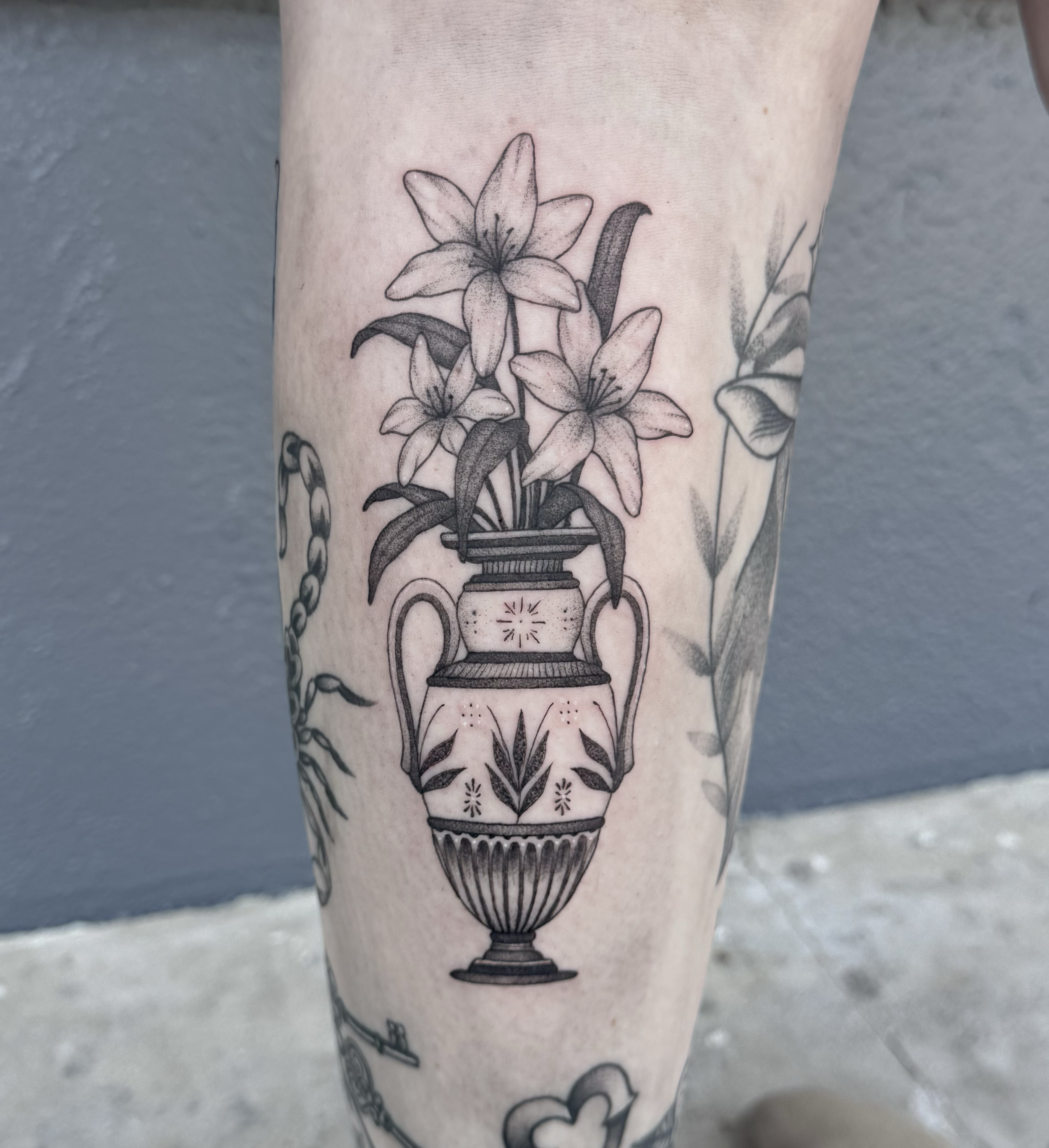 Black and gray tattoo of a bouquet of lilies in a decorative vase tattooed on a person's leg.