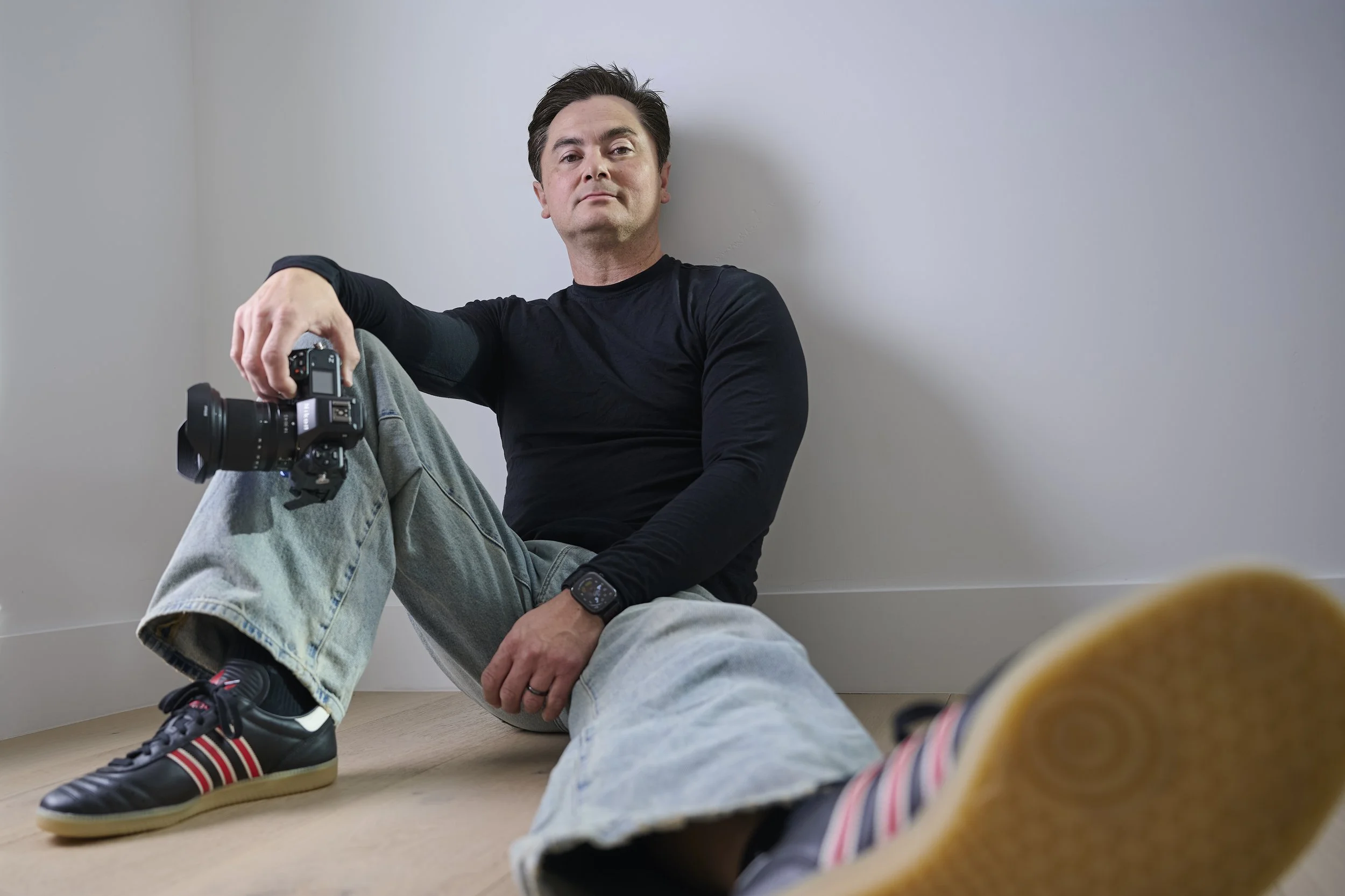 A man sitting on the floor against a white wall, holding a camera, wearing a black long-sleeve shirt, light gray jeans, and Adidas sneakers with red and white stripes, with a relaxed and confident expression.