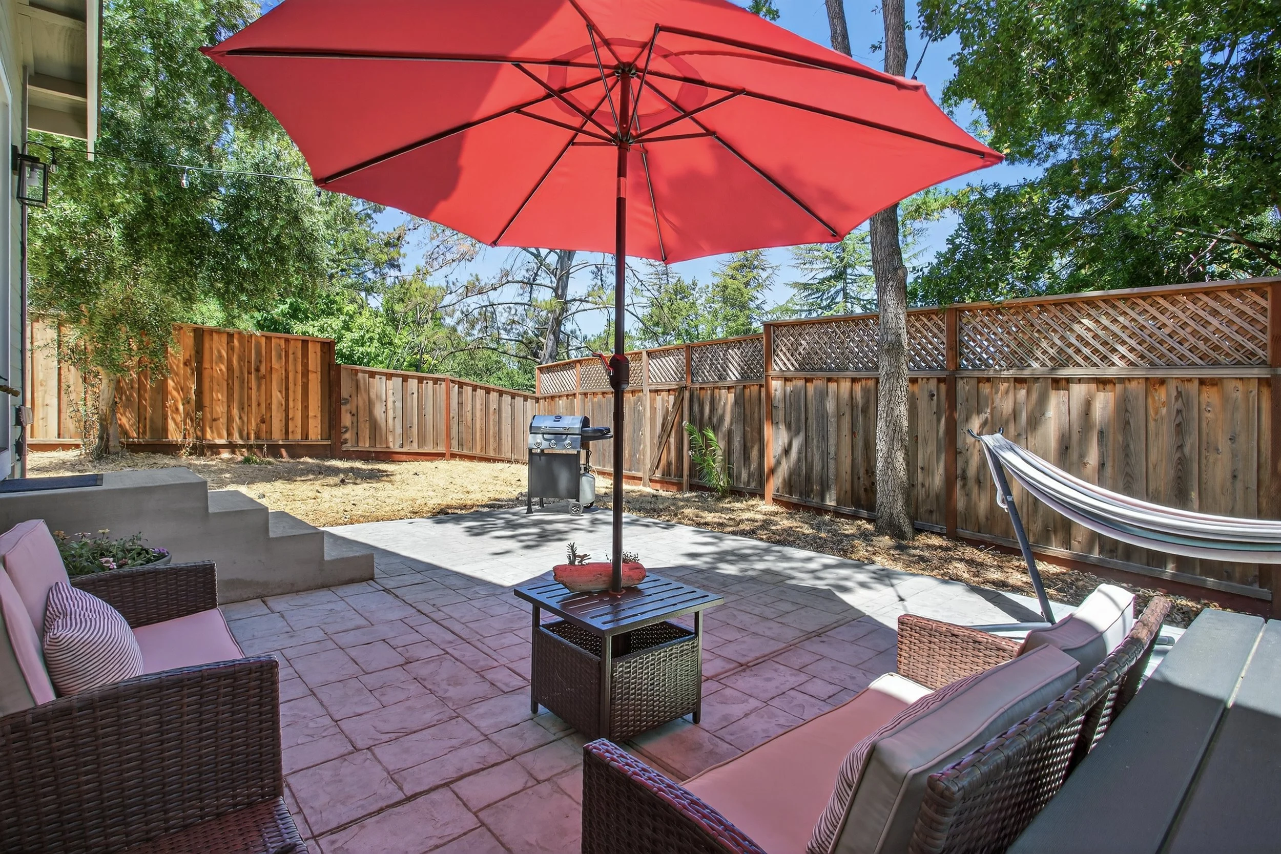 Backyard patio with outdoor furniture, large red patio umbrella, grill, hammock, wooden fence, trees, and blue sky.