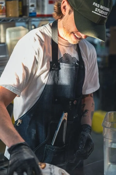 A man wearing a black cap and apron preparing food in a kitchen.