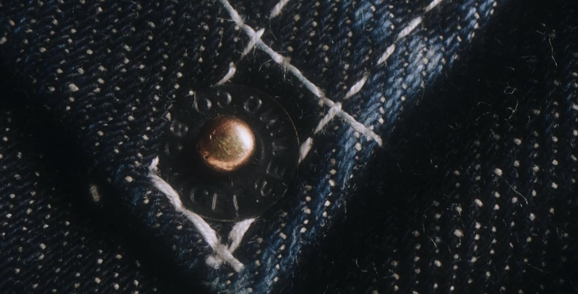 Close-up of a dark denim fabric with a metal button and white stitching.