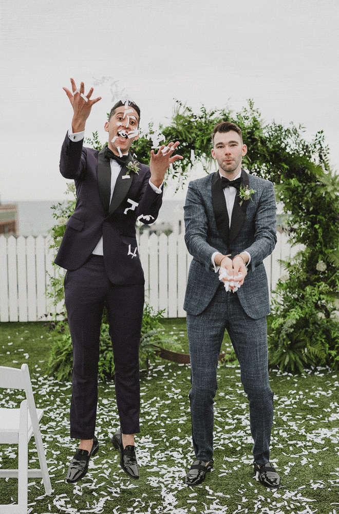 Grooms throwing confetti in front of lush greenery ceremony arch