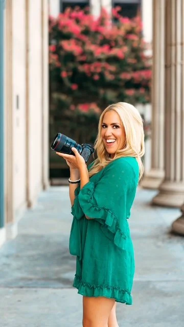 A smiling woman with blonde hair in a green dress holding a camera, standing on a walkway with columns and a flowering pink tree in the background.