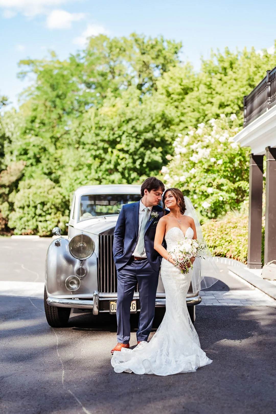 A bride and groom stand in front of a vintage silver car outdoors, with lush green trees in the background. The bride is wearing a white strapless wedding gown and holding a bouquet, while the groom is in a navy blue suit. They are closely gazing at each other, sharing an intimate moment.