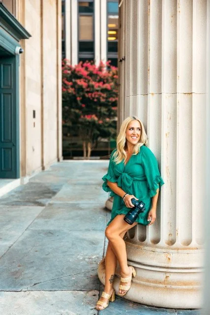 A woman with blonde hair in a turquoise dress sitting on a column outside a building, holding a camera, smiling, with pink flowers and buildings in the background.