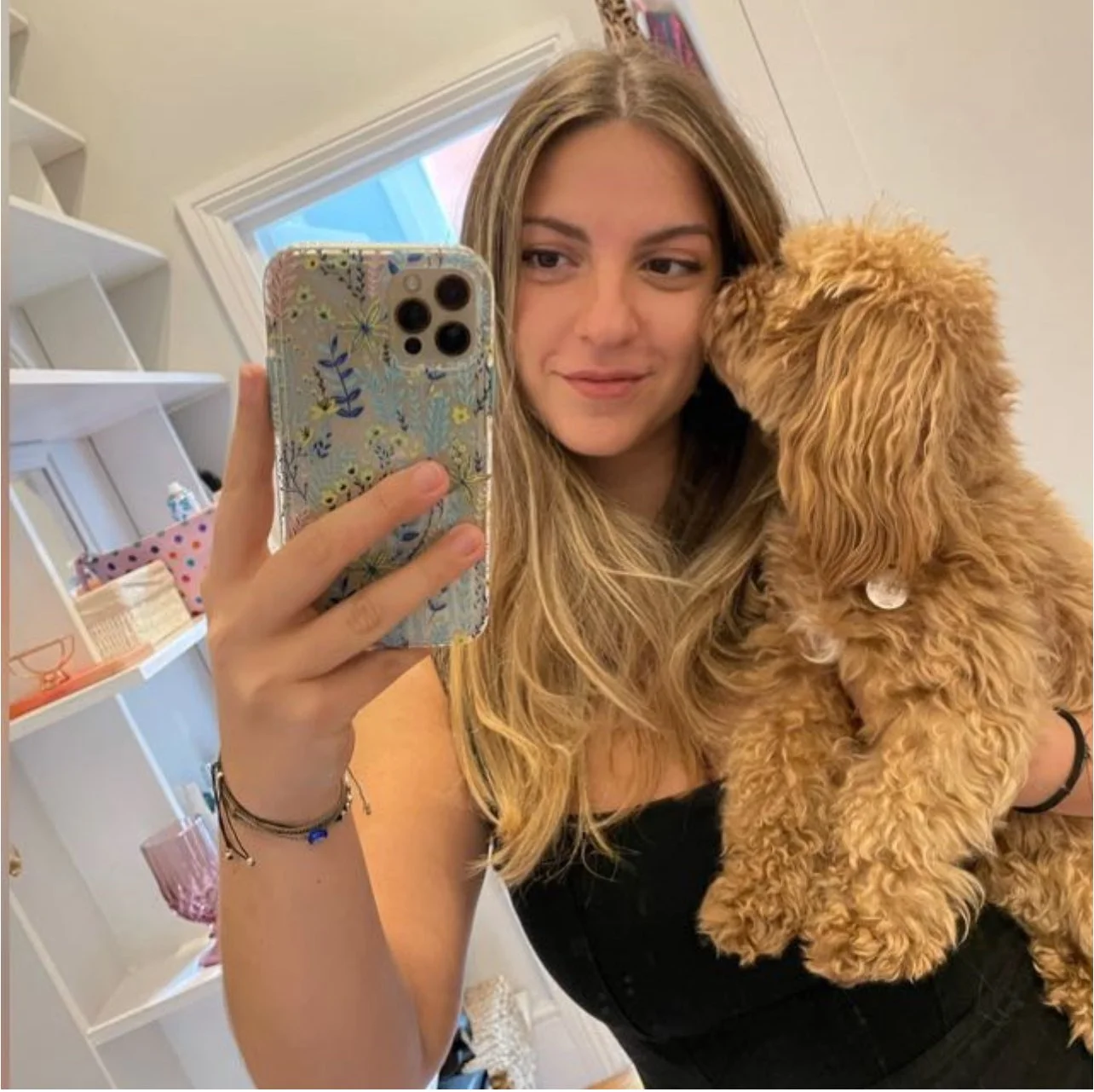 Young woman with long blonde hair taking a mirror selfie while holding a small curly-haired brown dog in front of a white background with shelves and a pink pillow.