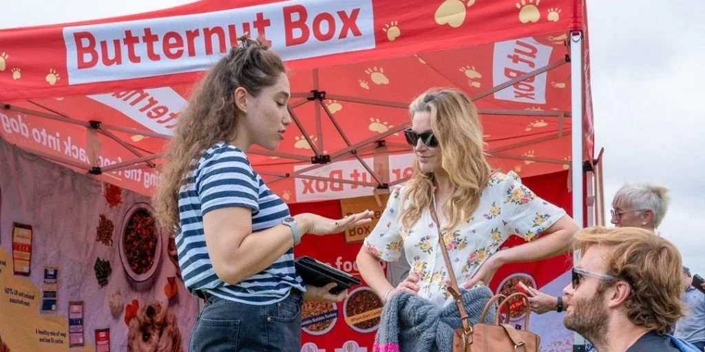 Three women and a man at a booth with a red canopy labeled 'Butternut Box'. The woman on the left, in a striped shirt, is showing something on her phone to the woman in glasses with wavy hair. The man with glasses and a beard, seated, looks towards the women. In the background, there is an older man with white hair.
