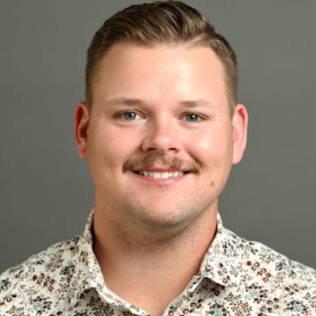 A young man with short, light brown hair, blue eyes, a light mustache, and a goatee, smiling in front of a gray background, wearing a floral-patterned shirt.