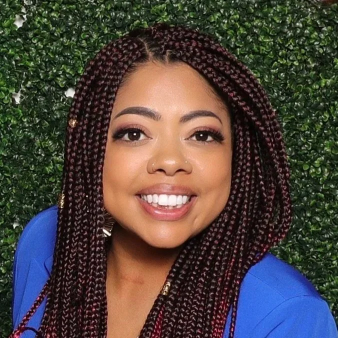 A woman with long, braided hair smiling in front of a green leafy background.
