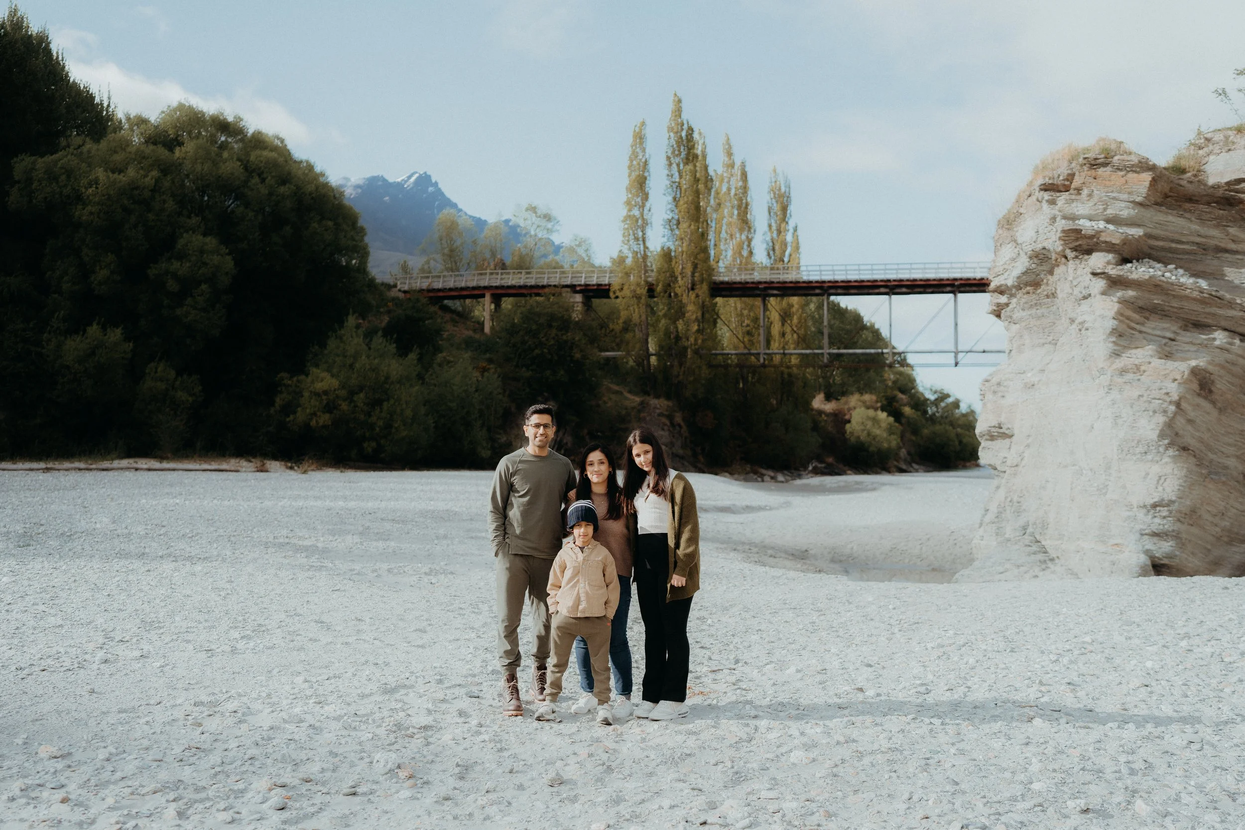 family of 4 posing in dry riverbed with the old shotover bridge in background