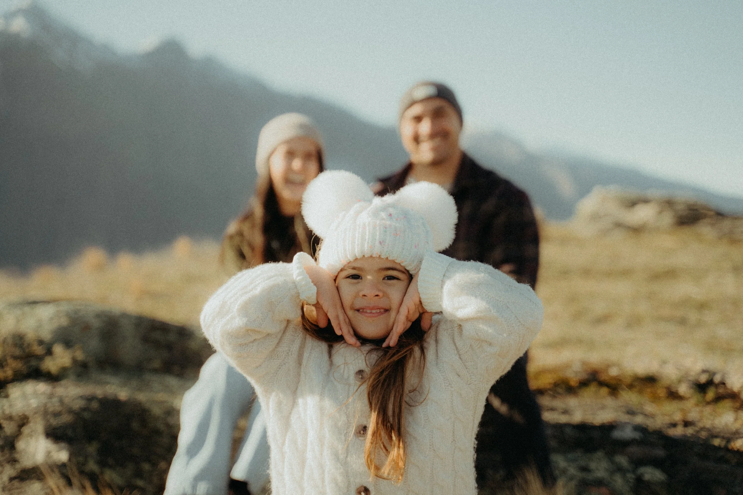 Girl smiling at camera with hands framing her chin, wearing a micky mouse beanie and parents smiling in background, with coronet peak behind them