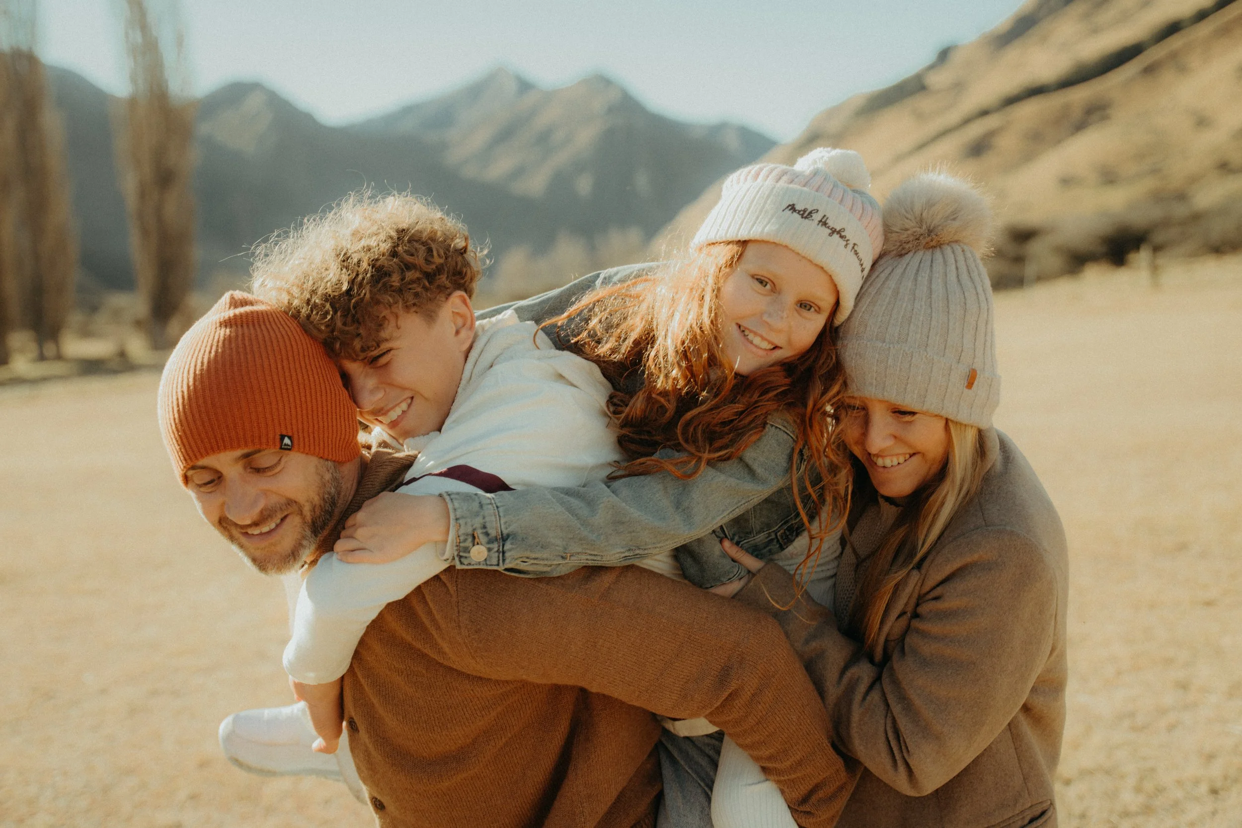 A family of 4 all hugging each other and smiling, surrounded by hills, dried grass and mountains at moke lake