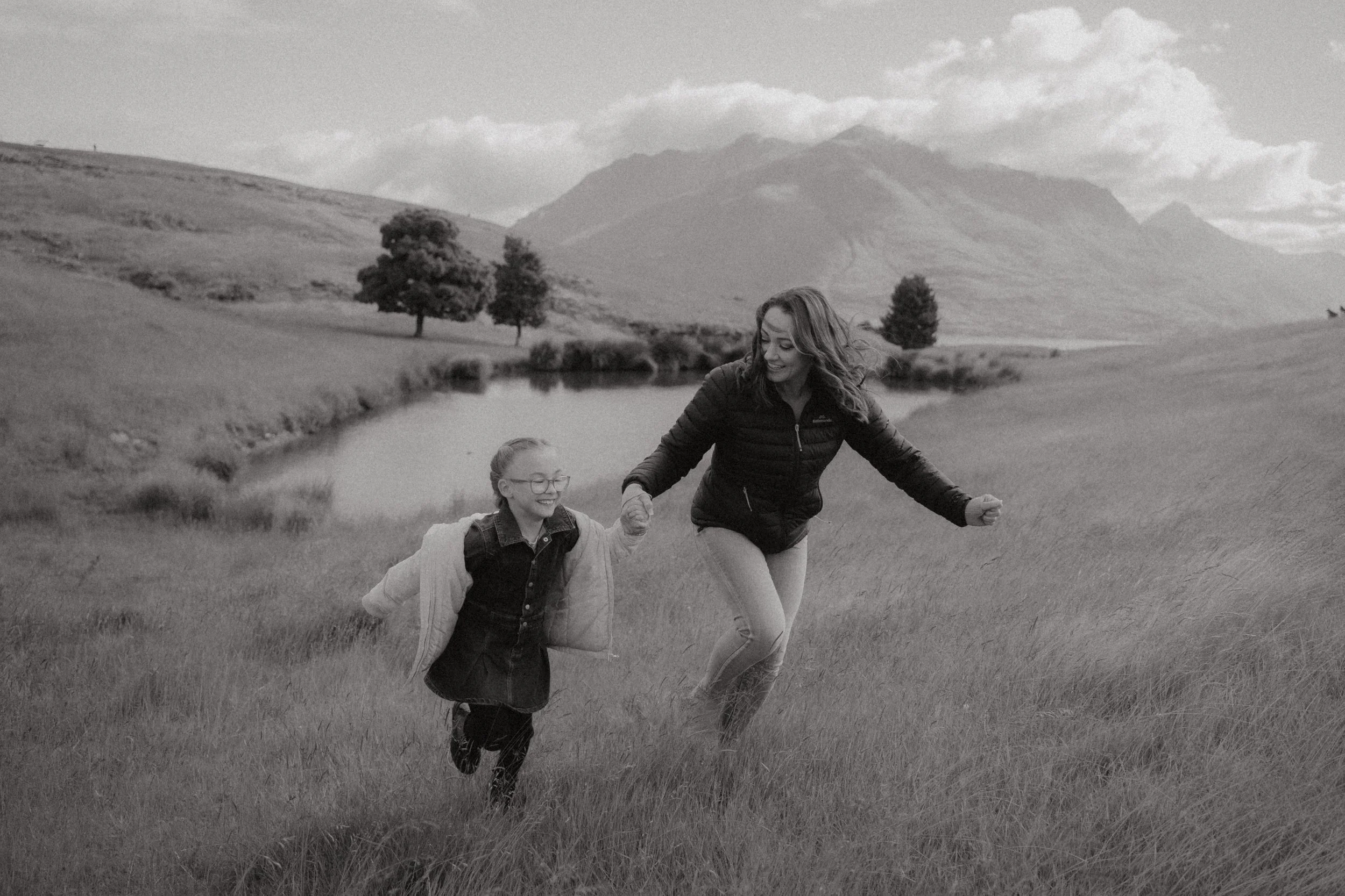 A  black and white image of a mother and a young daughter holding hands and running through a grassy field with a small pond, trees and rolling hills at deer park heights with coromandel peak mountains in the background during daytime.