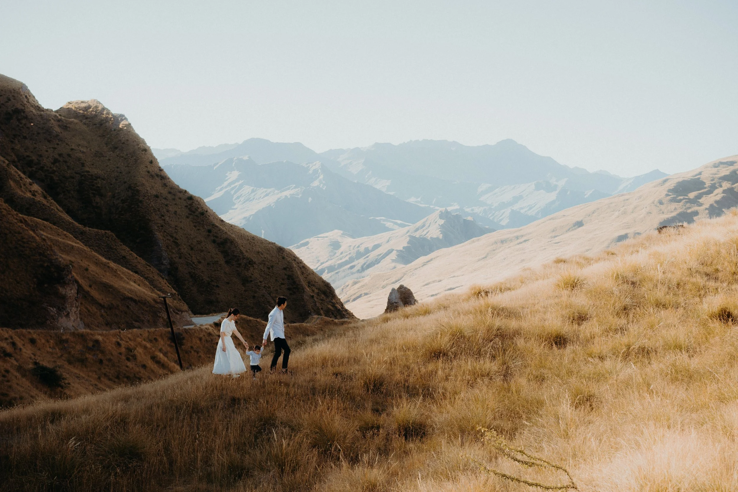 Family of 3 walking up grassy hill surrounded by mountains