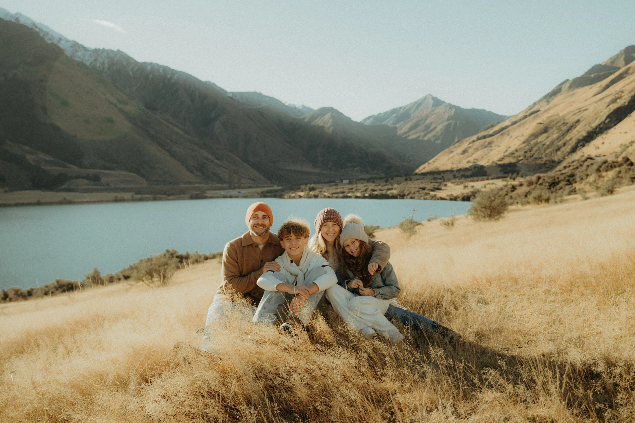 Family of 4 hugging each other on grassy field next to Moke Lake with mountains in the background on a sunny day..