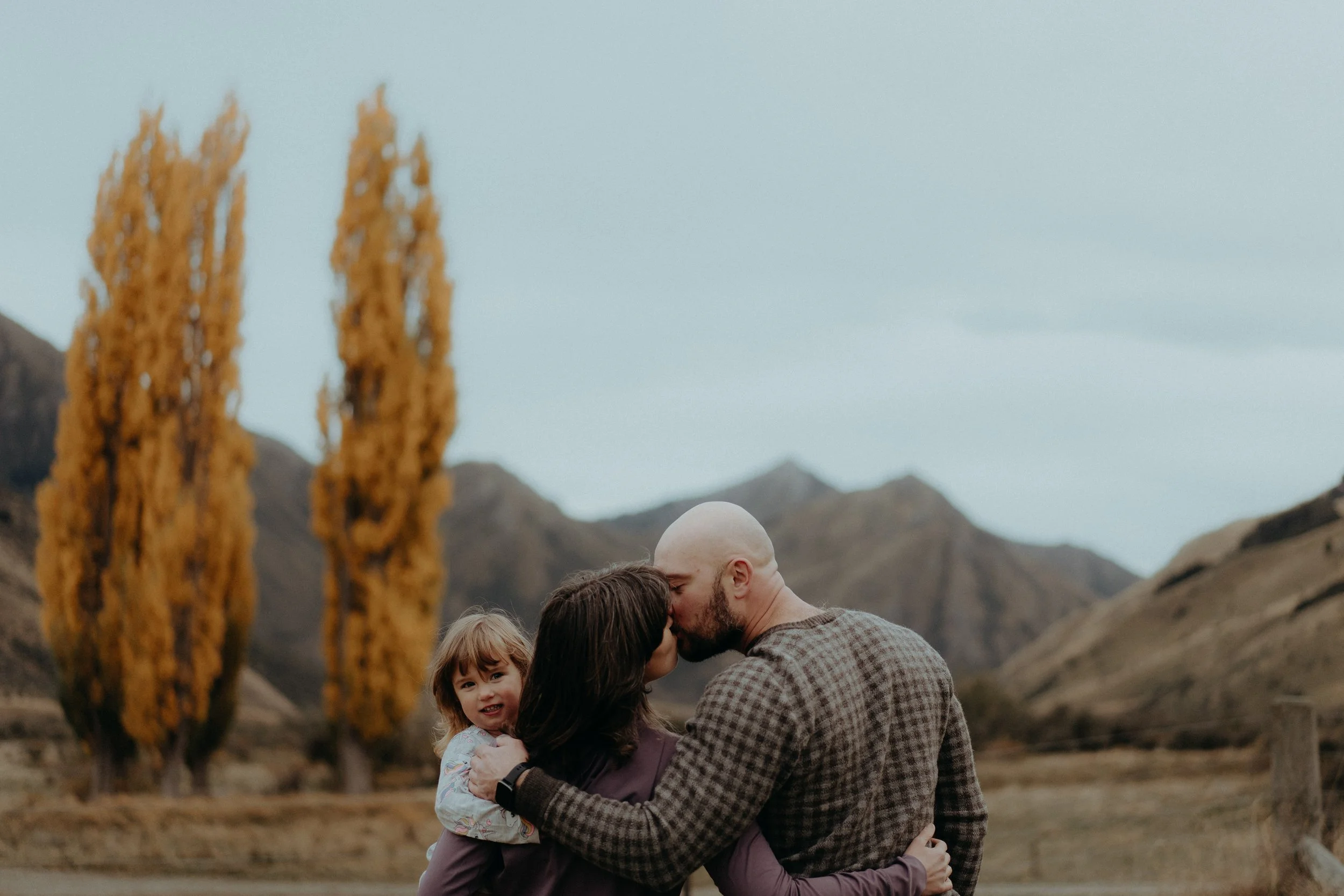 Family of 3 with parents kissing standing in front of autumn poplar trees