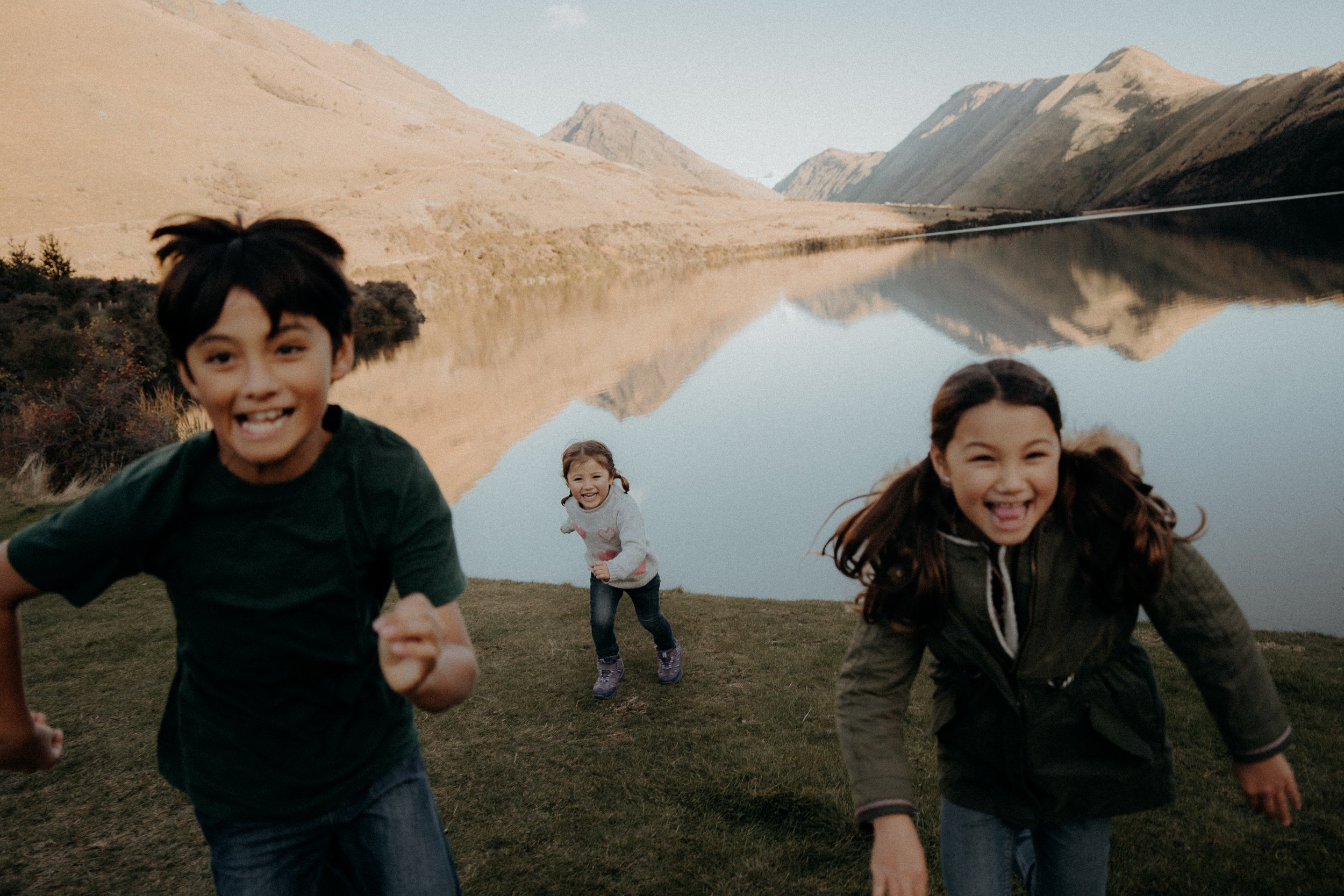 3 Kids playing tag, running away from their youngest sibling on the grassy shores of Moke Lake
