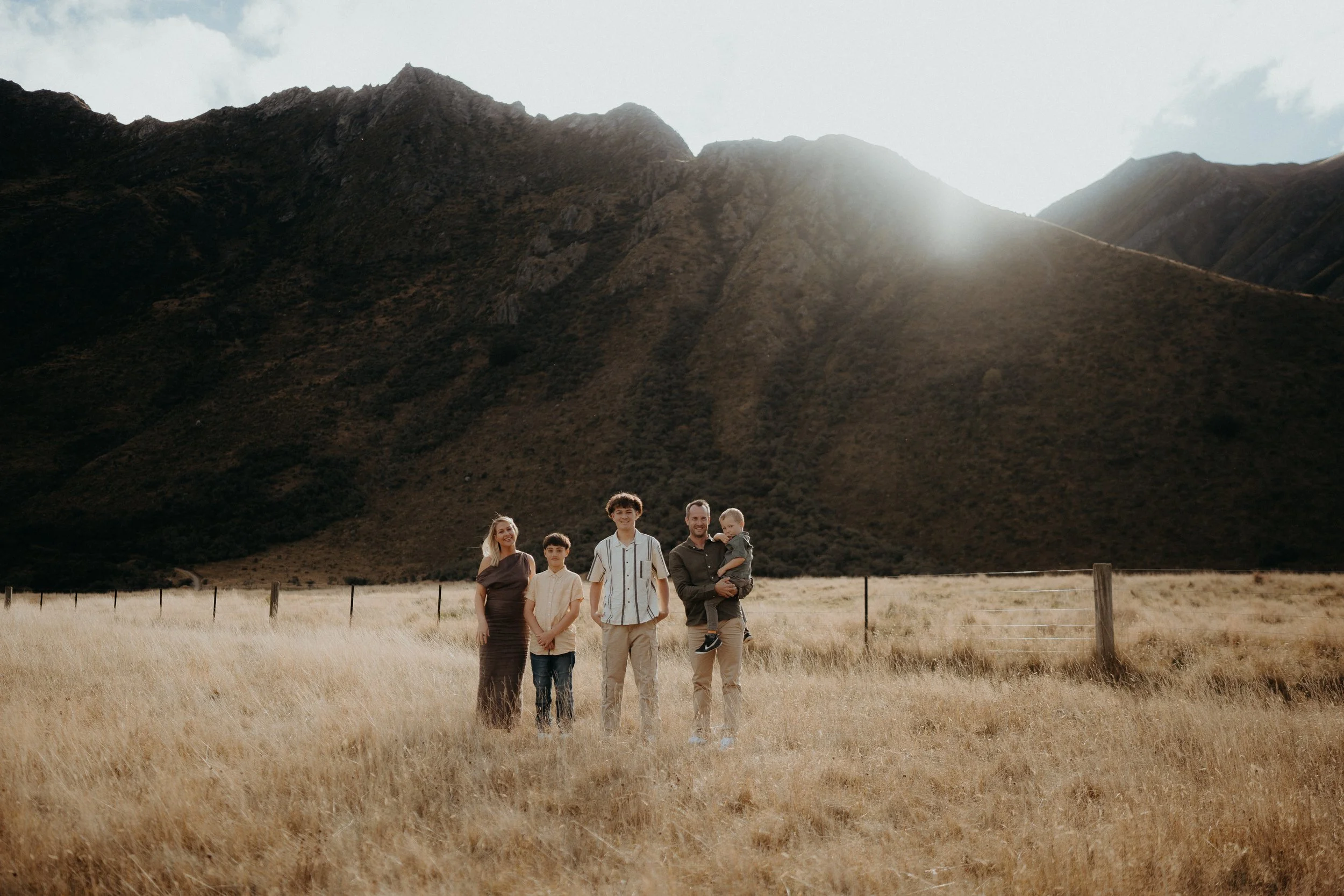 family of 5 standing in grassy field with sun going down behind a mountain