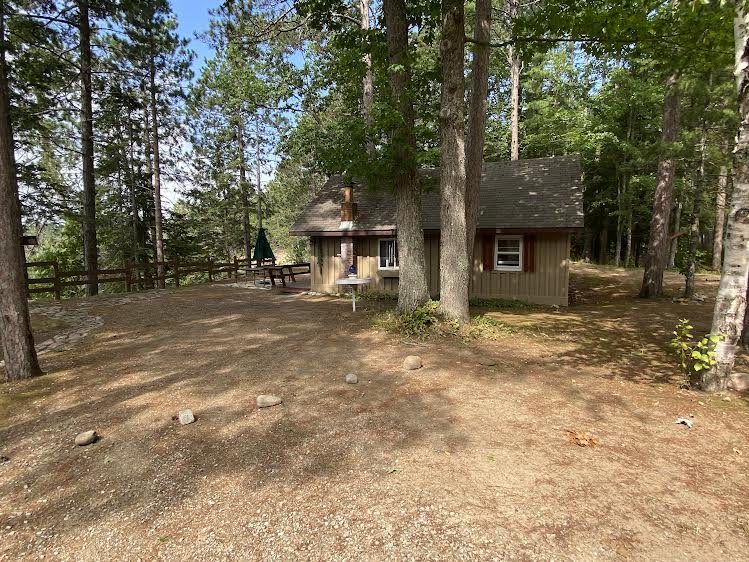 A small cabin in a wooded area with tall trees, a dirt ground, and a picnic table with an umbrella outside.