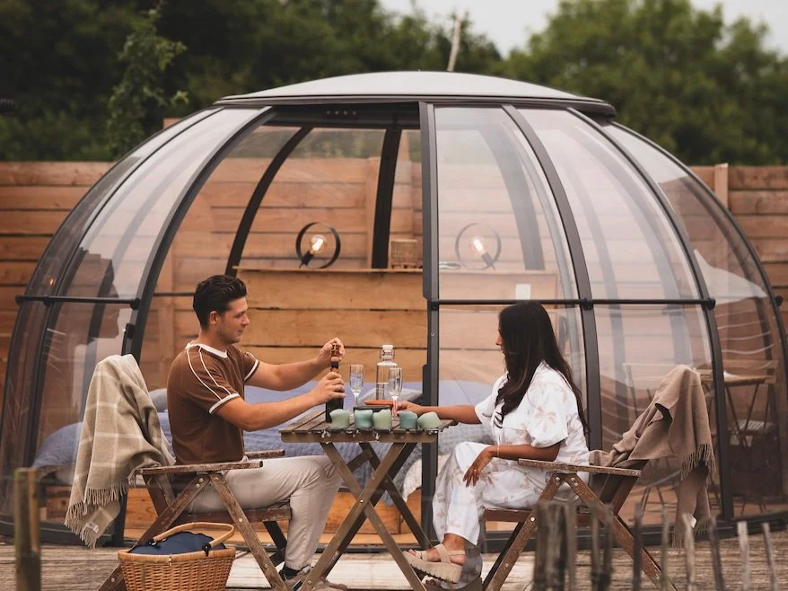 Un couple assis à une table d'extérieur, tenant un toast, dans une serre en verre avec un fond de bois.