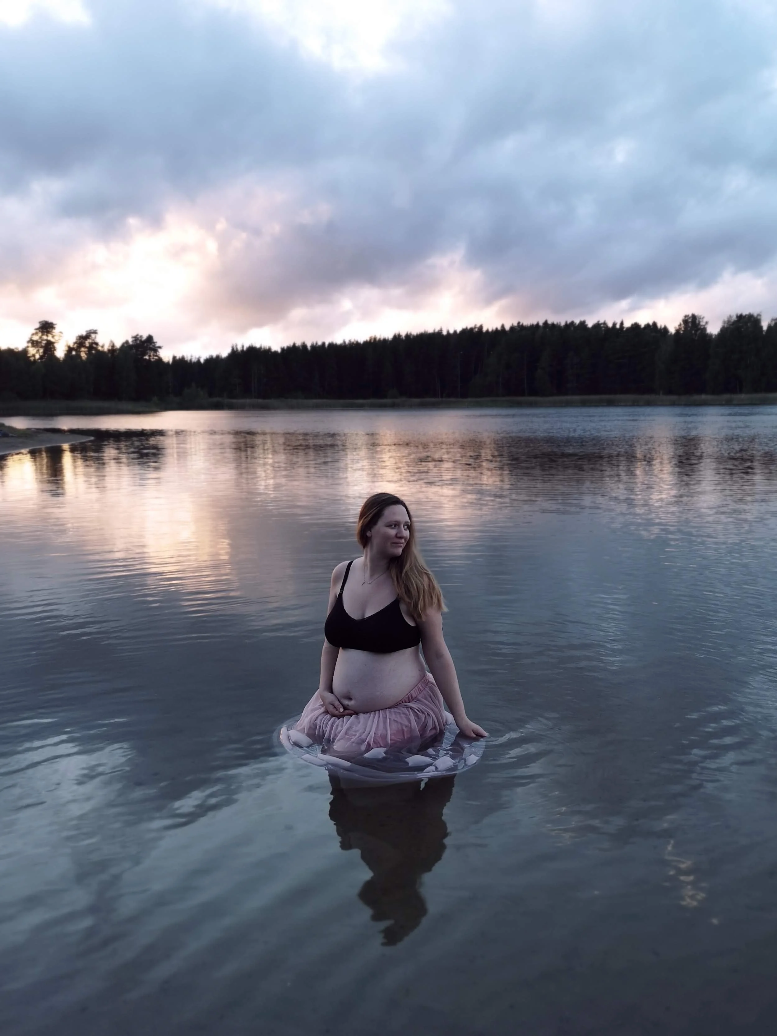 En gravid kvinna som bär en svart topp och en rosa kjol står i vattnet vid en sjö under en molnig himmel.