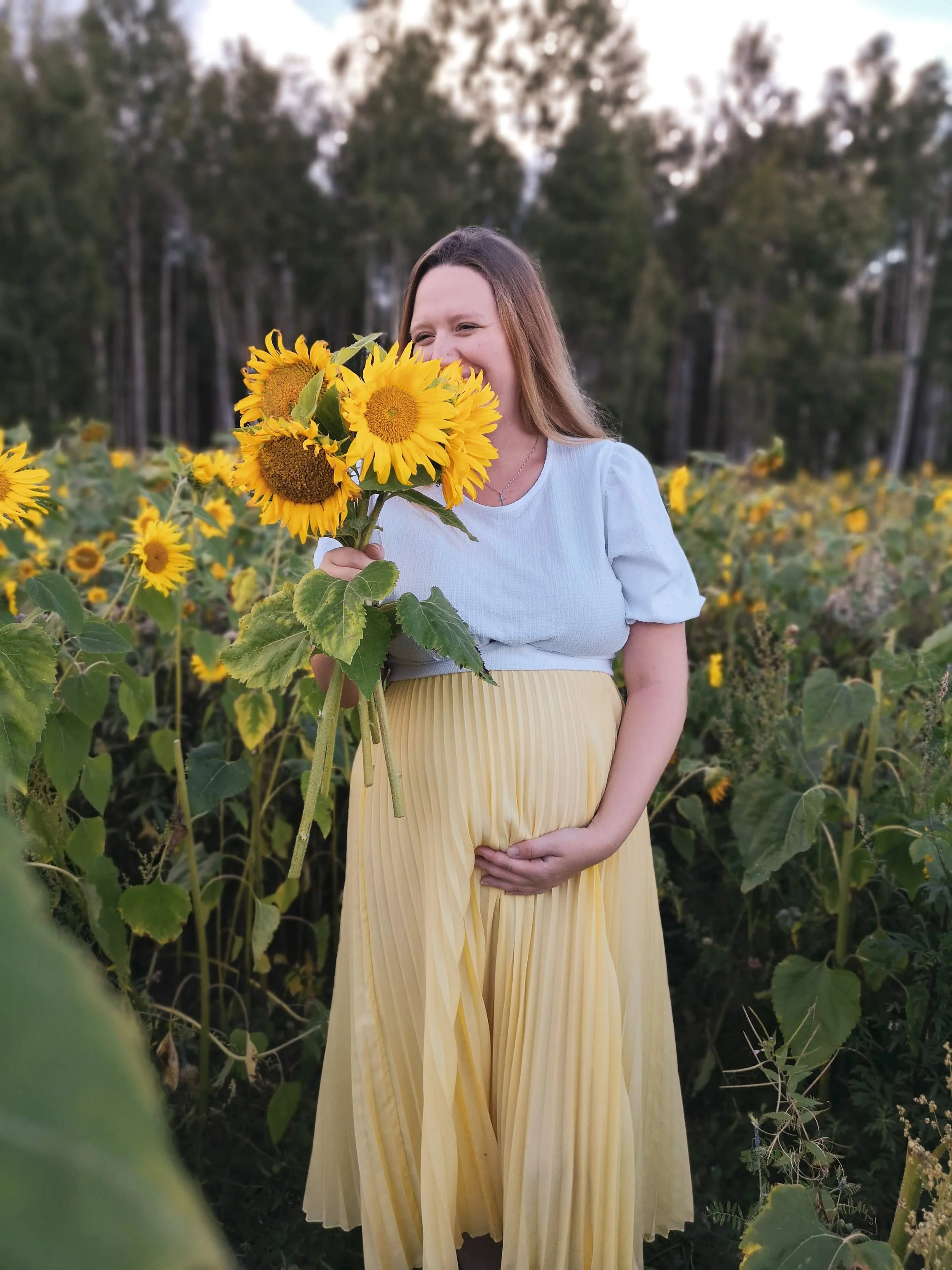 Ung gravid  kvinna i en blomsteräng håller en bukett solrosor framför ansiktet, bär ljusblått och gult, med skog i bakgrunden.