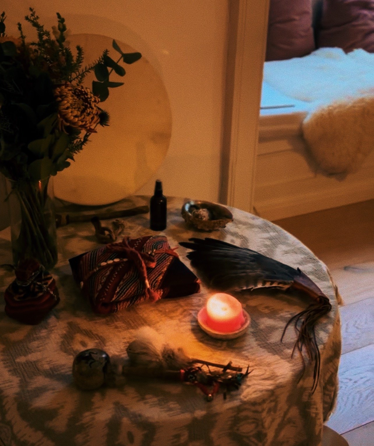 A table with a bouquet of flowers, a wrapped package, a feather, a small bottle, a candle, and decorative stones, with a window in the background.