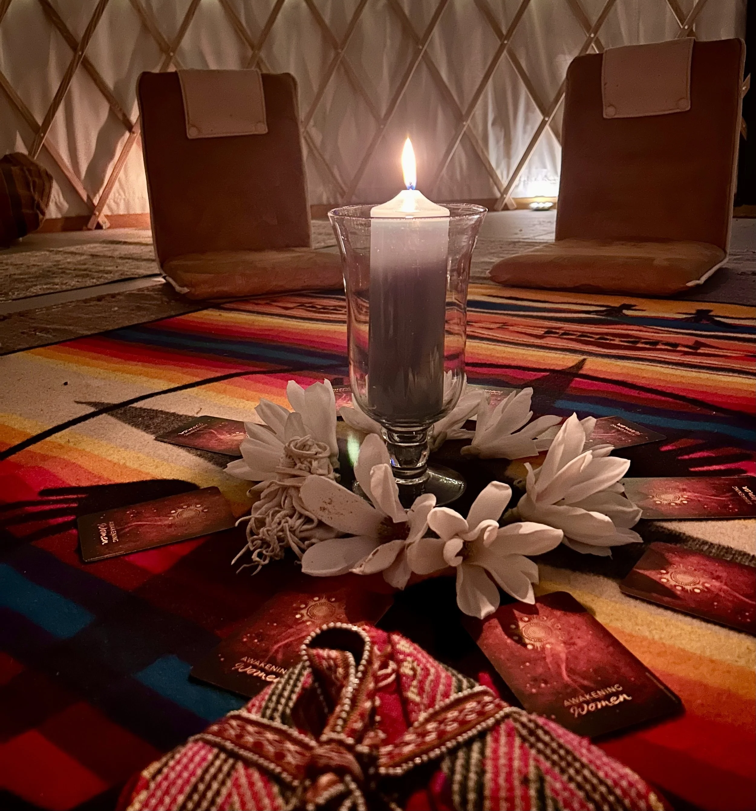 A wooden table with a colorful tribal patterned tablecloth, surrounded by two brown chairs, features a lit white candle in a clear glass holder, with white flower petals and tarot cards arranged in a circle around the candle.