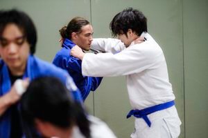 Two judo practitioners, one in a blue gi and the other in a white gi, engaged in a match with focus and intensity, while another person in a blue gi is blurred in the foreground.