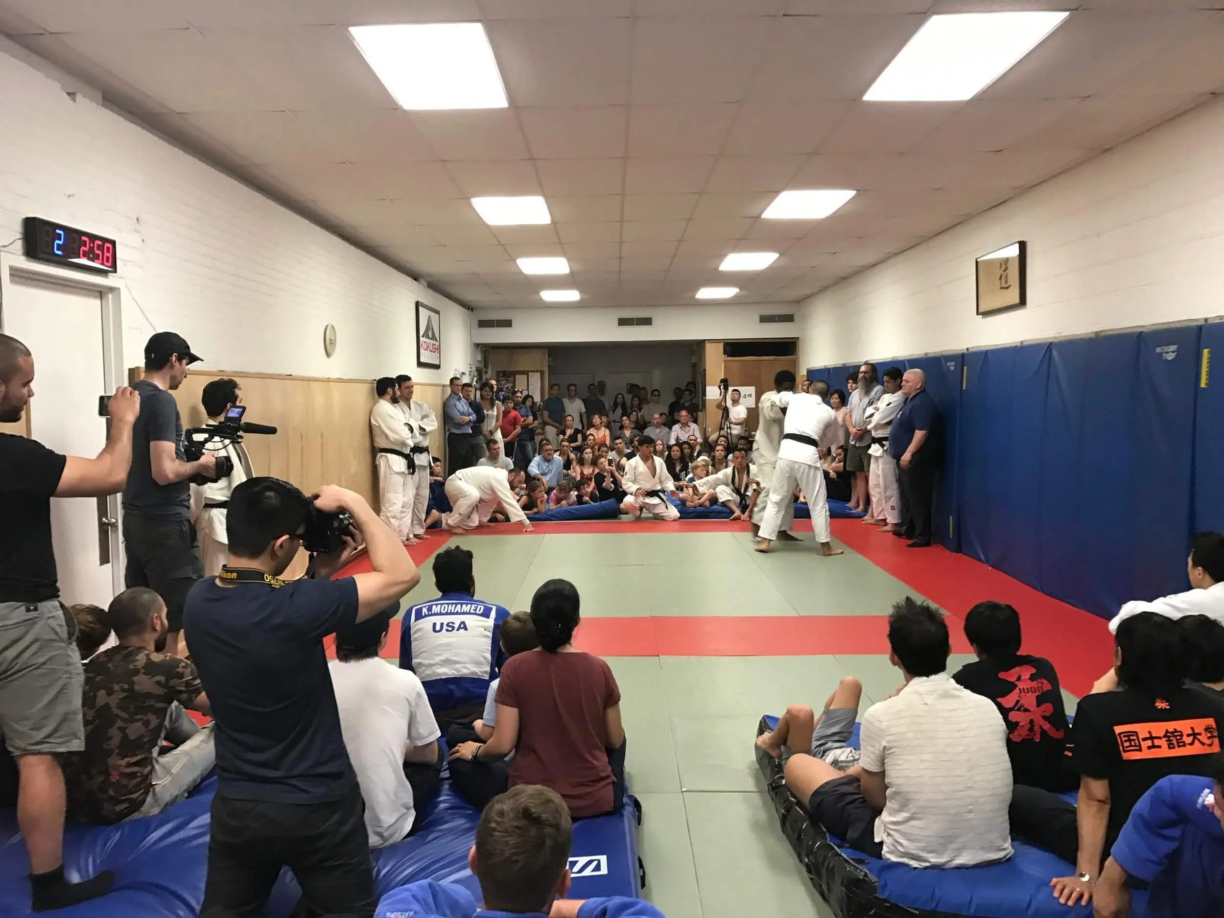 Martial arts training session or competition with people sitting and standing around a matted training area, some practicing techniques, and a large audience watching. Several individuals in white gis, one with a black belt, are in the center, while 