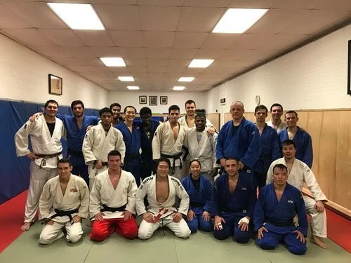 Group photo of Judo practitioners in a dojo, wearing judo gis, some kneeling and others standing, with a white wall, framed pictures, and a clock in the background.