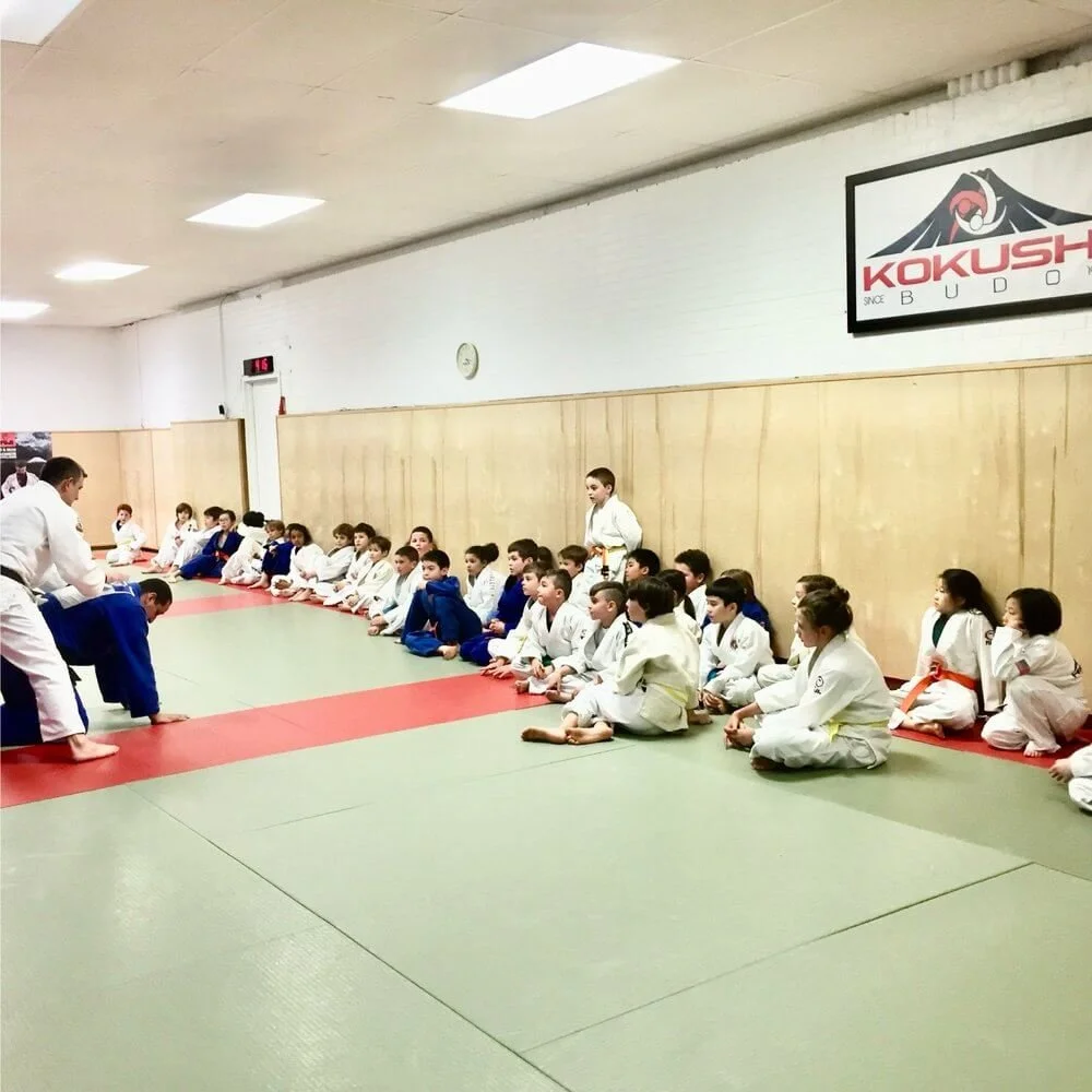 Children sitting on the mat while a martial arts instructor demonstrates techniques in a dojo. The children are wearing gis and colored belts, and the dojo has a wooden panel wall with a large logo of 'KOKUSHI' and 'BUDO' on a banner.