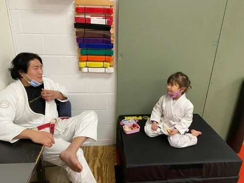 A man and a young girl sit facing each other in a room, both in white martial arts uniforms, wearing masks, sharing a light moment. The girl is sitting on a black mat with a pink lunchbox beside her, and the man is sitting on a chair.