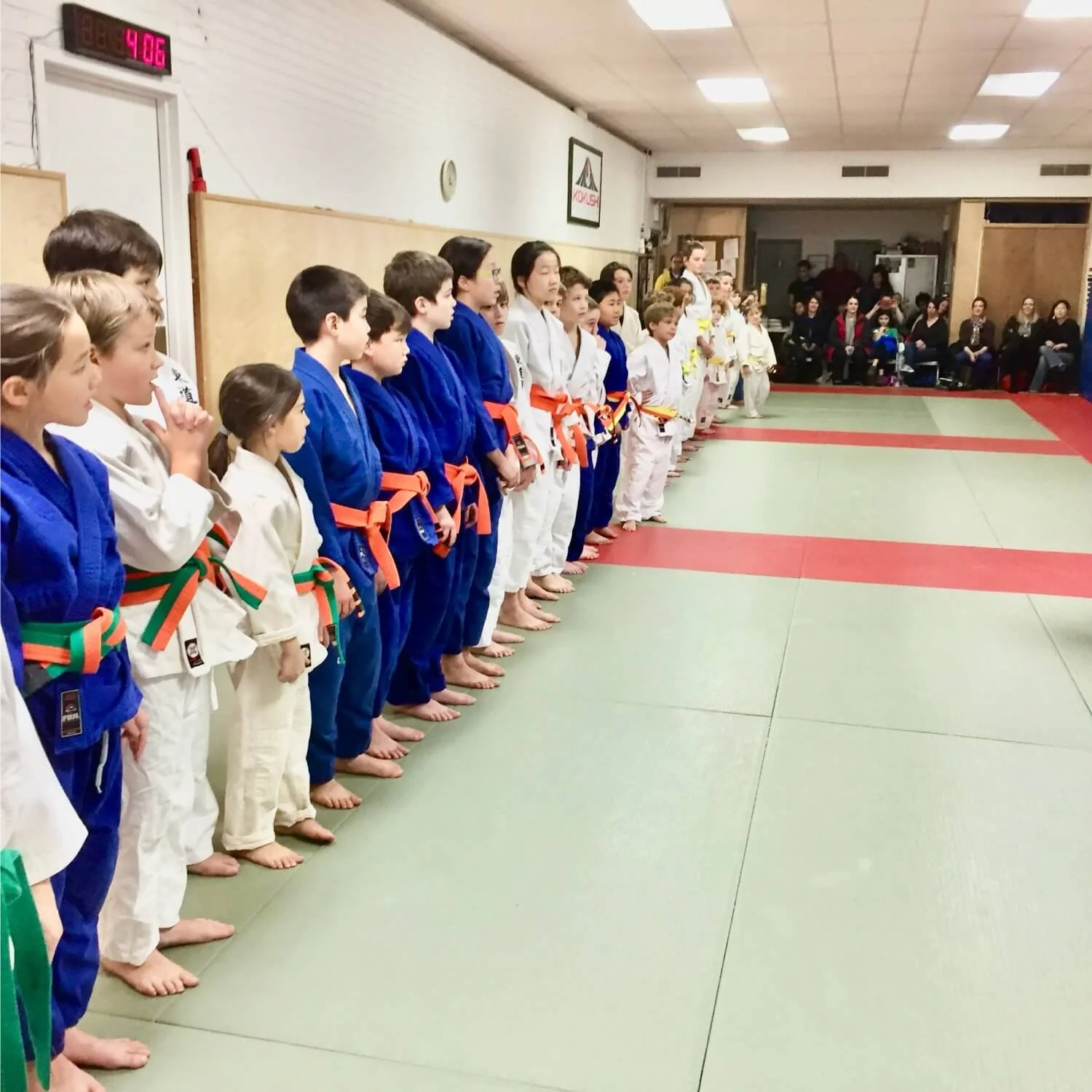 Children in judo uniforms standing in a line on judo mats during a ceremony or competition, with spectators seated in the background.