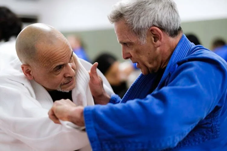 Two men practicing martial arts, one in a white gi, the other in a blue gi, engaged in a close combat technique in a dojo or training hall.
