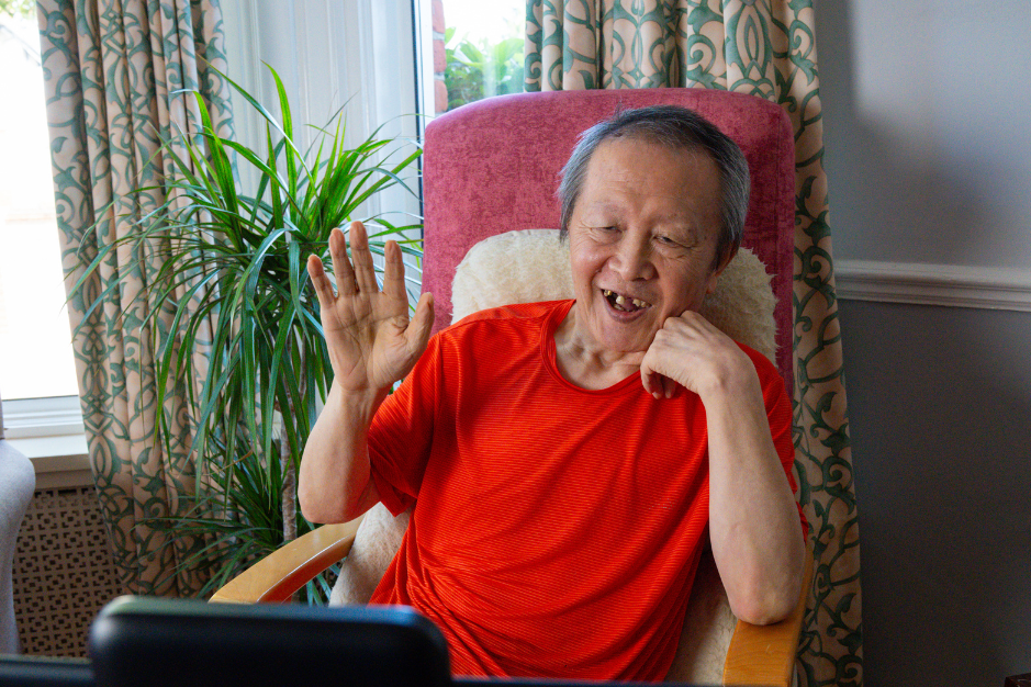An elderly man with a missing tooth, wearing a red shirt, sitting on a pink armchair, smiling and waving at a tablet or screen in front of him.