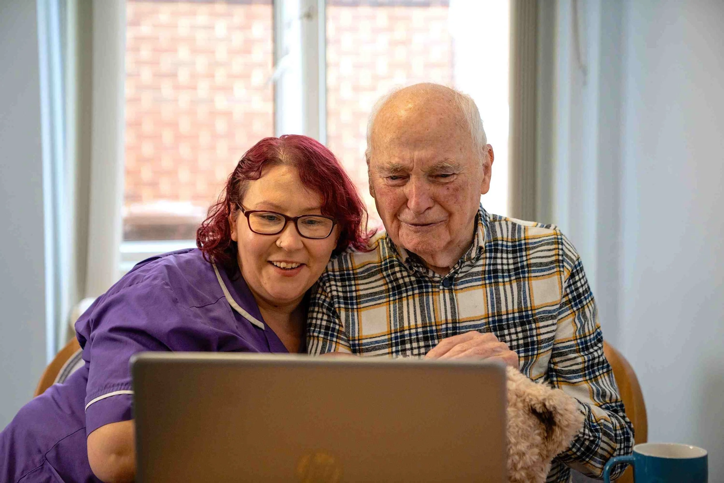 A woman with red hair and glasses in a purple uniform sitting next to an elderly man with a checkered shirt and a dog, looking at a laptop screen together in a bright room with large windows.