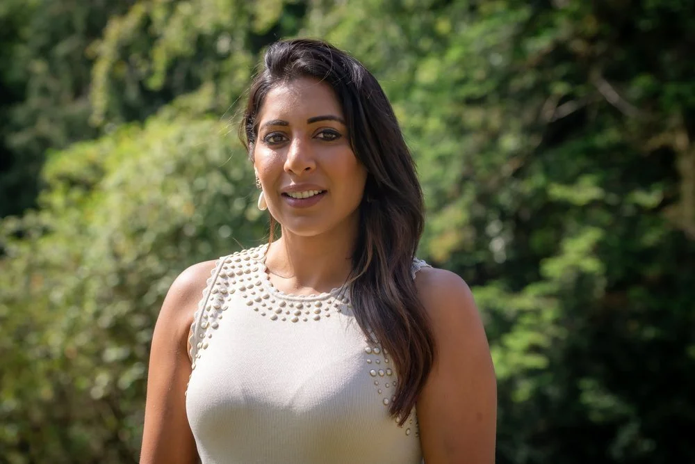 A woman with long dark hair standing outdoors in front of green trees sunlight.