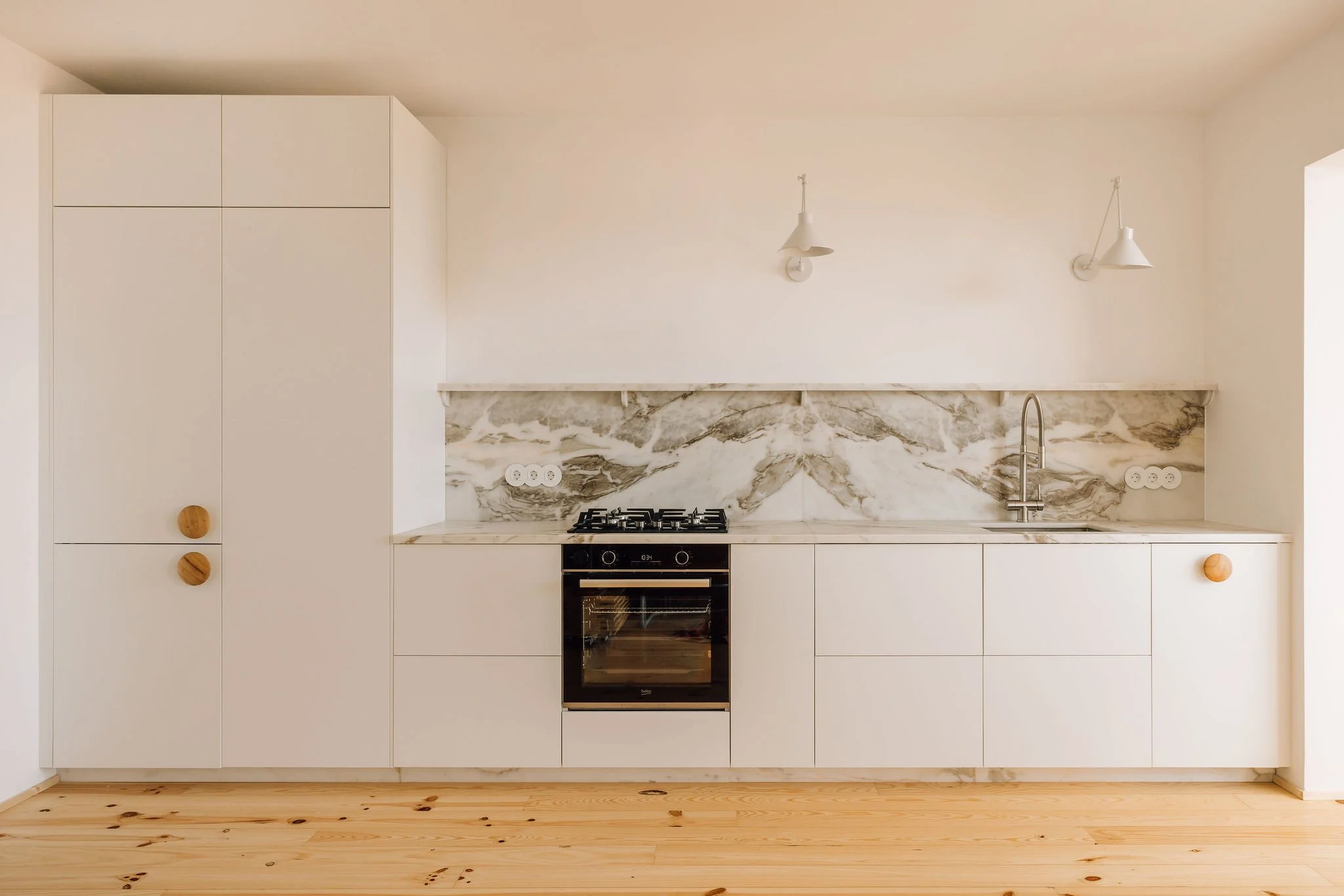 Minimalist kitchen with white cabinets, marble backsplash, built-in oven, stovetop, stainless steel sink, and wooden knobs, on light wooden flooring.