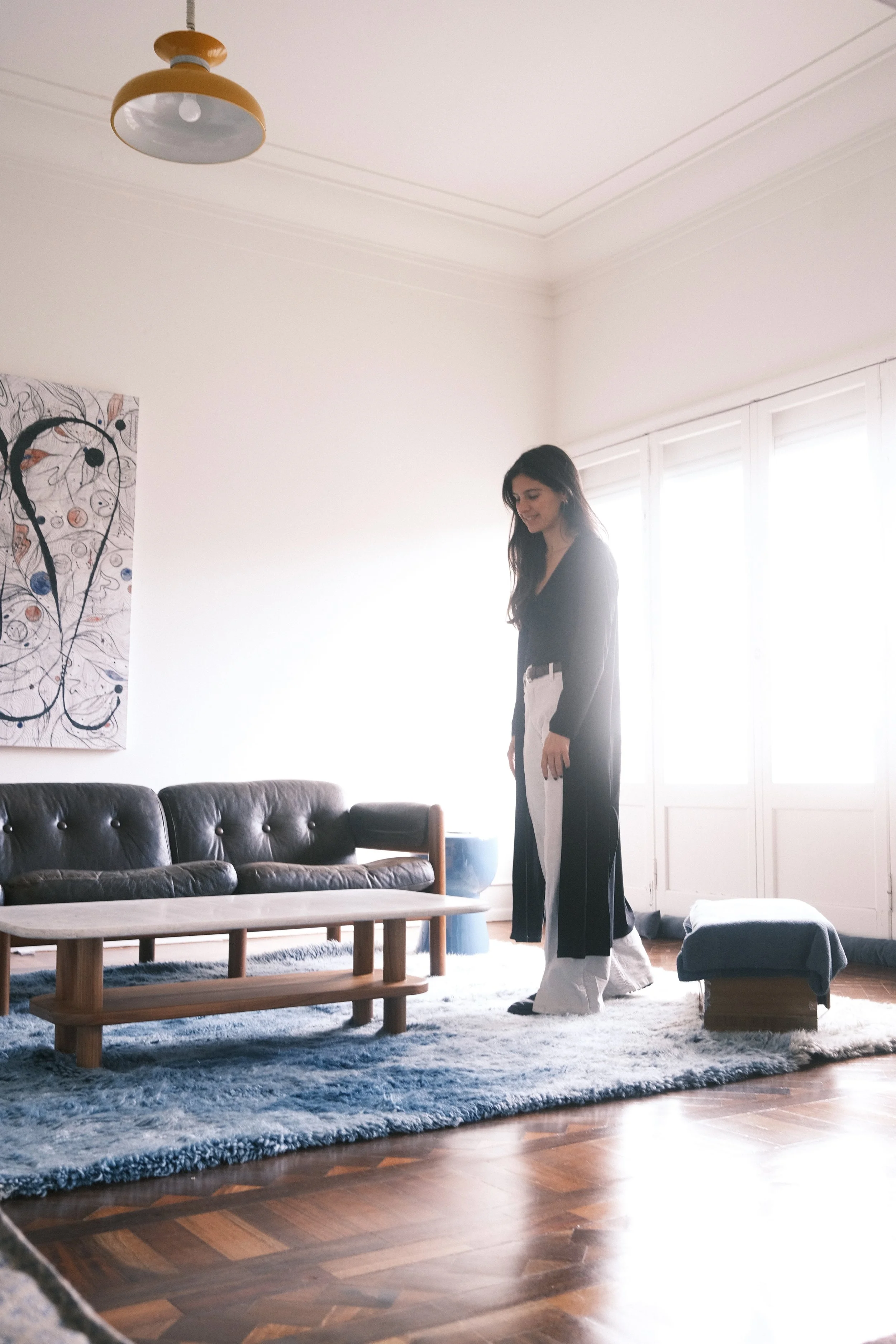 A woman standing in a living room with a dark leather sofa, a wooden coffee table, a blue and white rug, and large windows with bright sunlight.