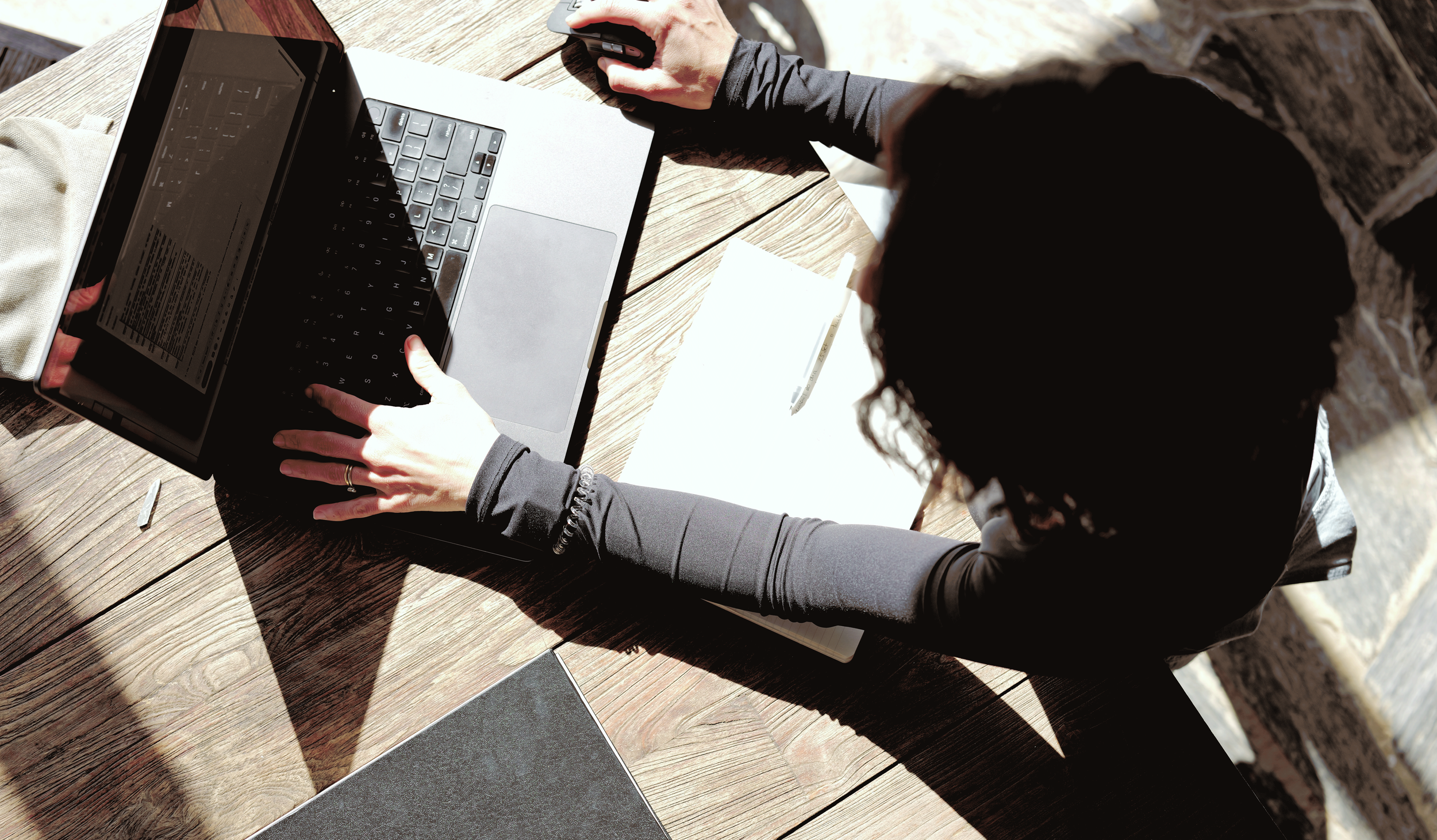 A person sitting at a wooden table using a laptop with a black keyboard and a black screen. They are wearing a black long-sleeve top, holding a computer mouse with one hand, and the other hand is on the laptop. There's a notepad and pen on the table, with sunlight casting shadows.