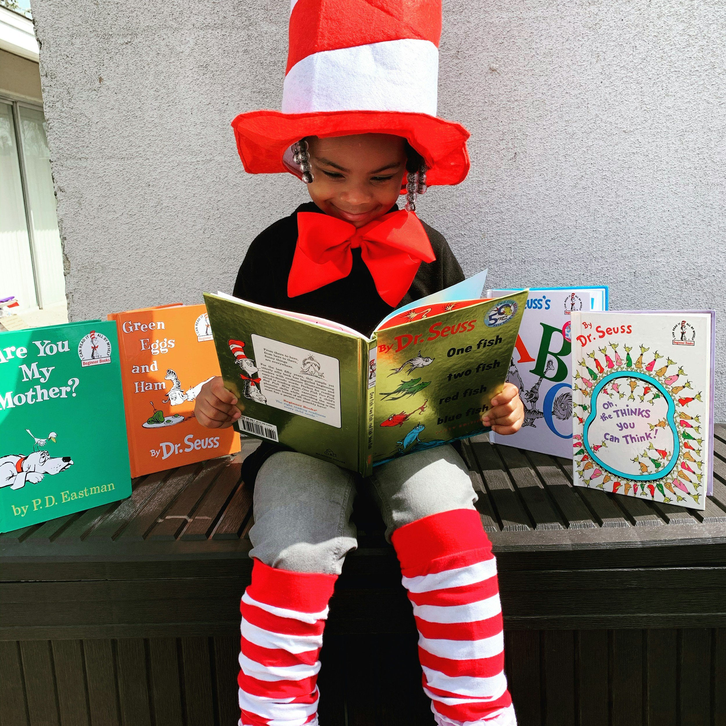 A young child dressed as the Cat in the Hat, wearing a tall red and white striped hat, a red bow tie, and red and white striped pants, reading a book outdoors near a gray wall.