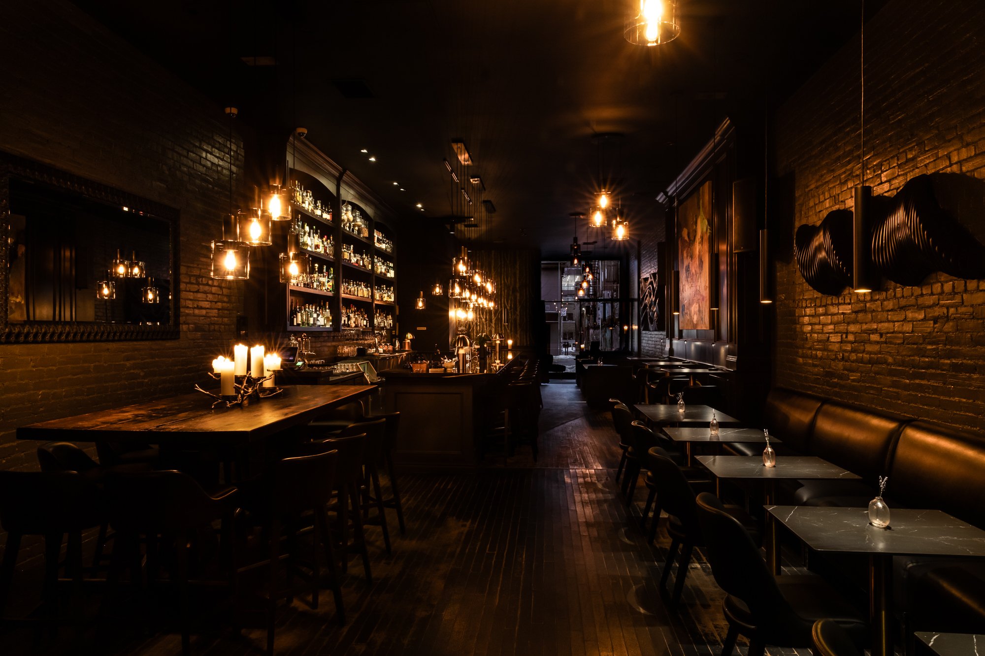 A cozy, dimly lit bar with a large selection of bottles on shelves, dark furniture, candles, and artwork on exposed brick walls.
